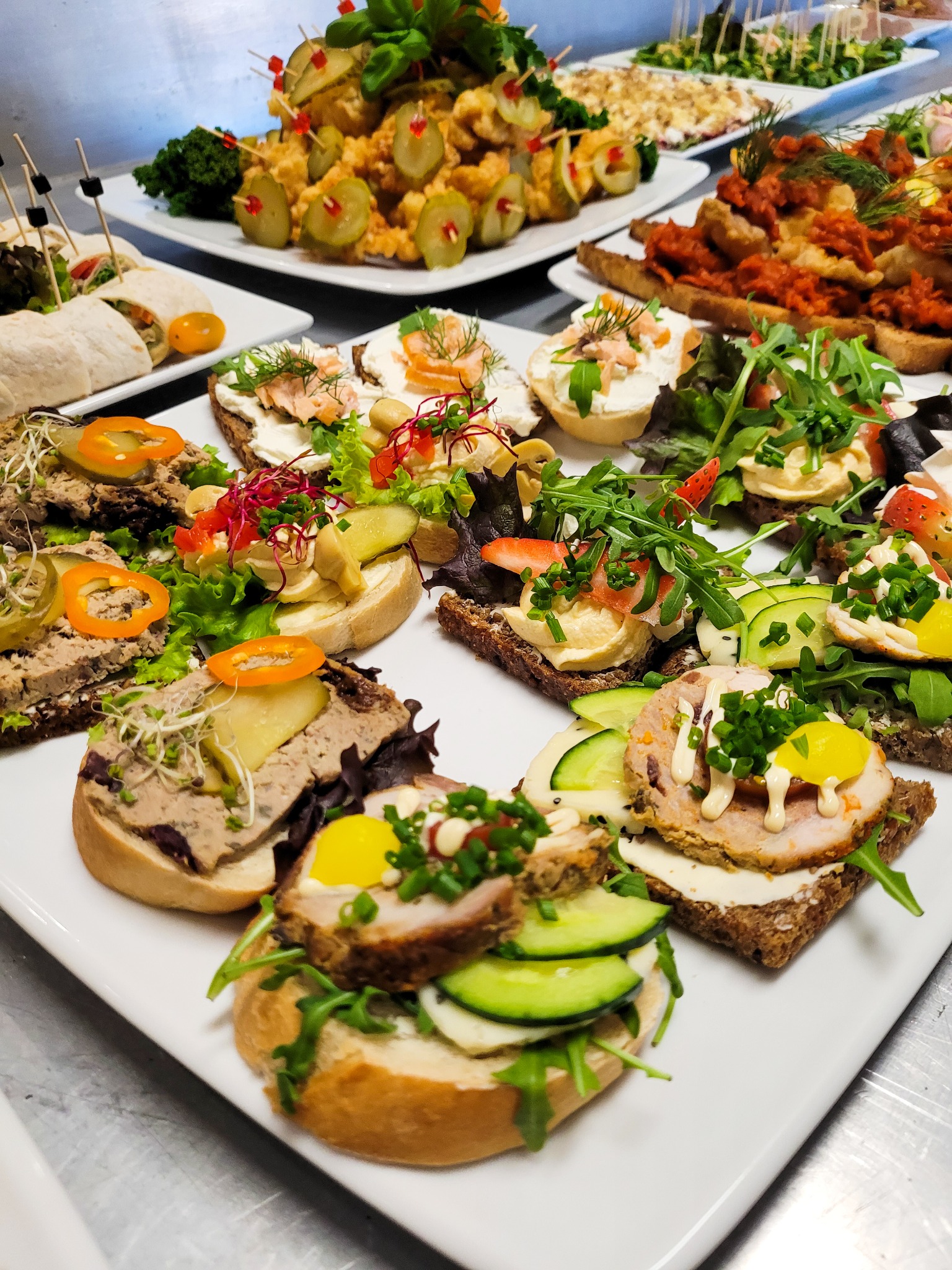 Różnorodne kanapki i przekąski na białych talerzach, w tym z pasztetem, łososiem, warzywami i owocami, ułożone na metalowym stole, przygotowane do serwowania.