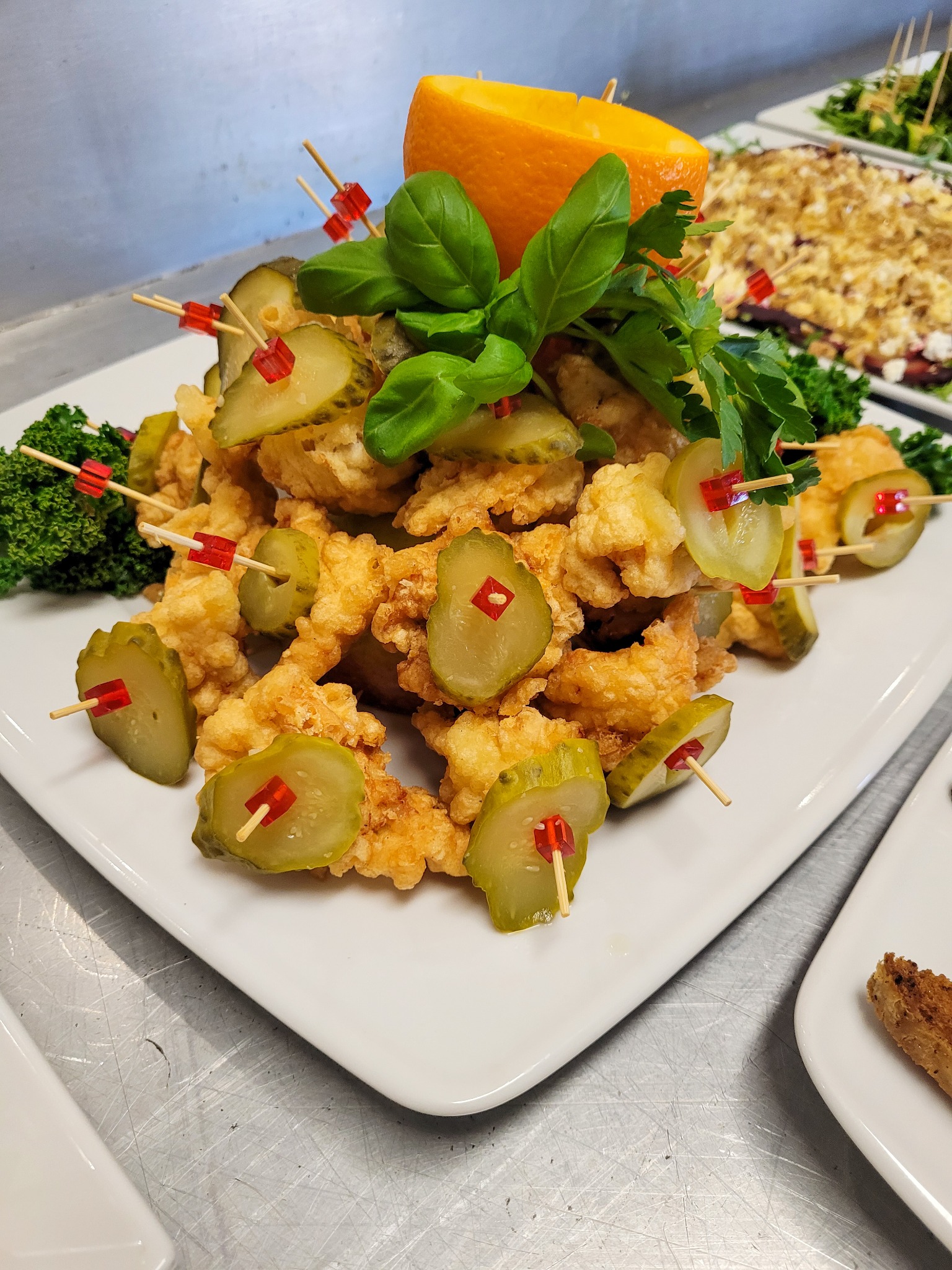 Biały talerz z przekąską: smażone kawałki kurczaka dekorowane plastrami ogórka konserwowego na wykałaczkach z czerwonymi ozdobami, bazylią, natką pietruszki i połówką pomarańczy. W tle inne potrawy...