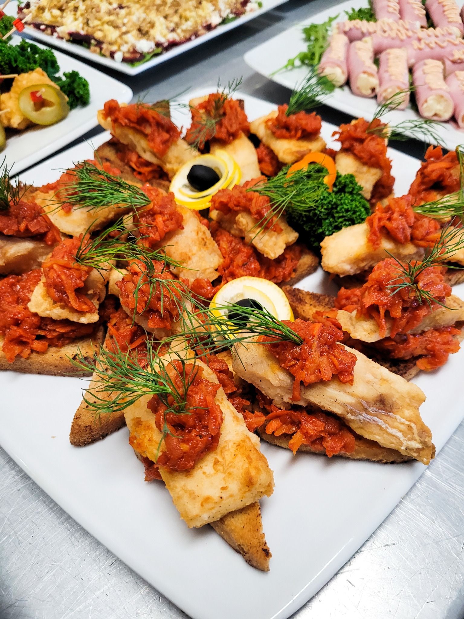 Elegancko ułożone na białym talerzu kanapeczki z rybą w panierce, czerwoną pastą warzywną i koperkiem, obok inne przekąski.