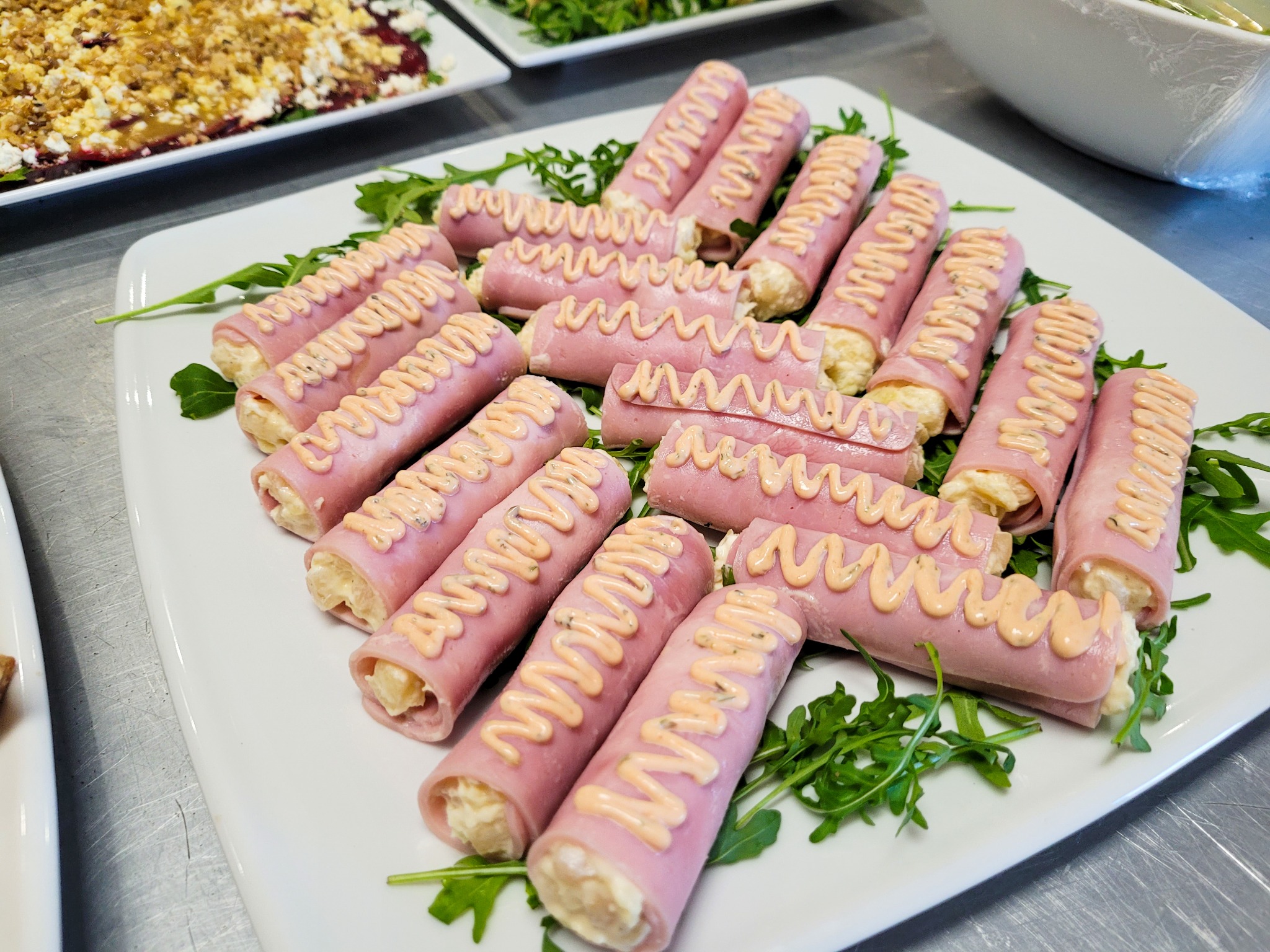 Roladki z szynki nadziewane sałatką, udekorowane sosem i rukolą na białym talerzu.