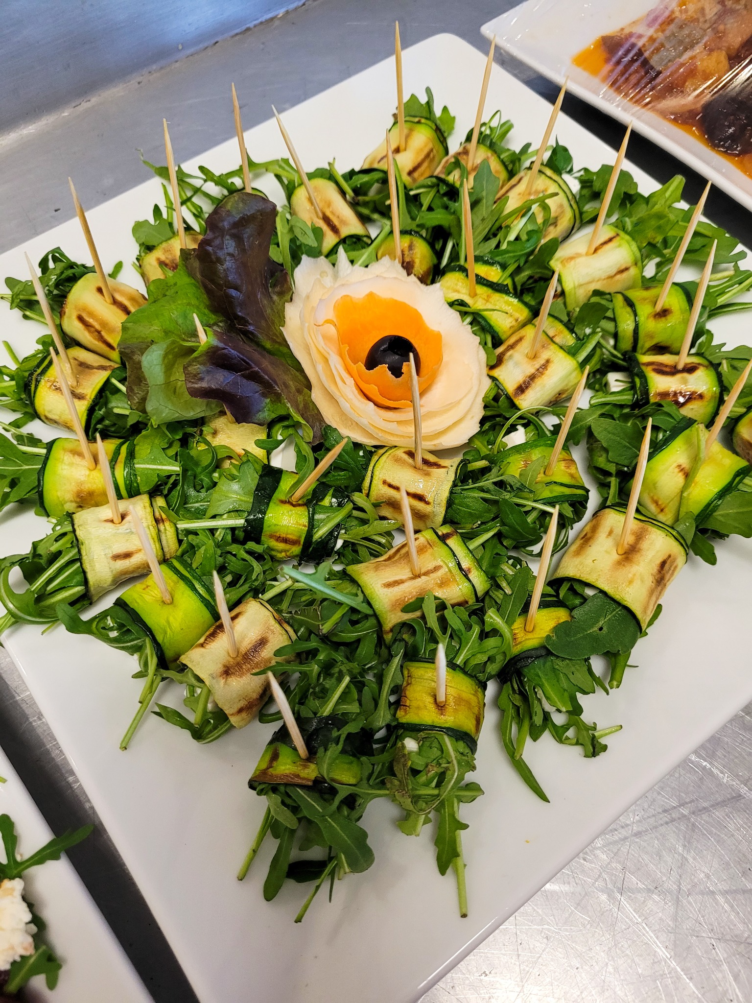 Elegancka kompozycja z grillowanej cukinii zwiniętej w ruloniki, spiętej wykałaczkami i ułożonej na świeżej rukoli, z dekoracyjnym elementem przypominającym kwiat z pomarańczowym środkiem,...