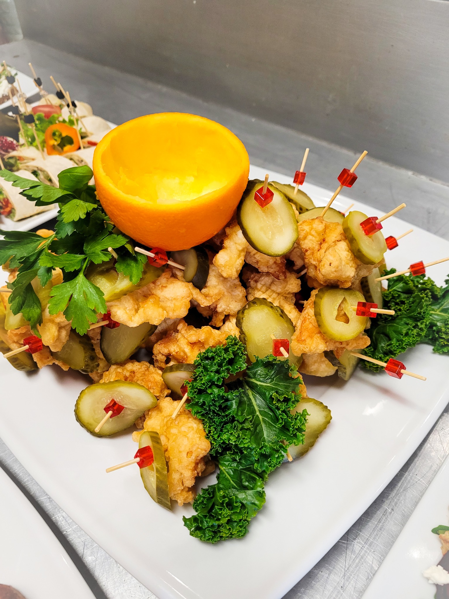 Dekoracyjna kompozycja z kawałków smażonego kurczaka i plastrów ogórka konserwowego na szpilkach z czerwonymi ozdobami, z połową pomarańczy i zieloną pietruszką, na białym talerzu. W tle widoczne...