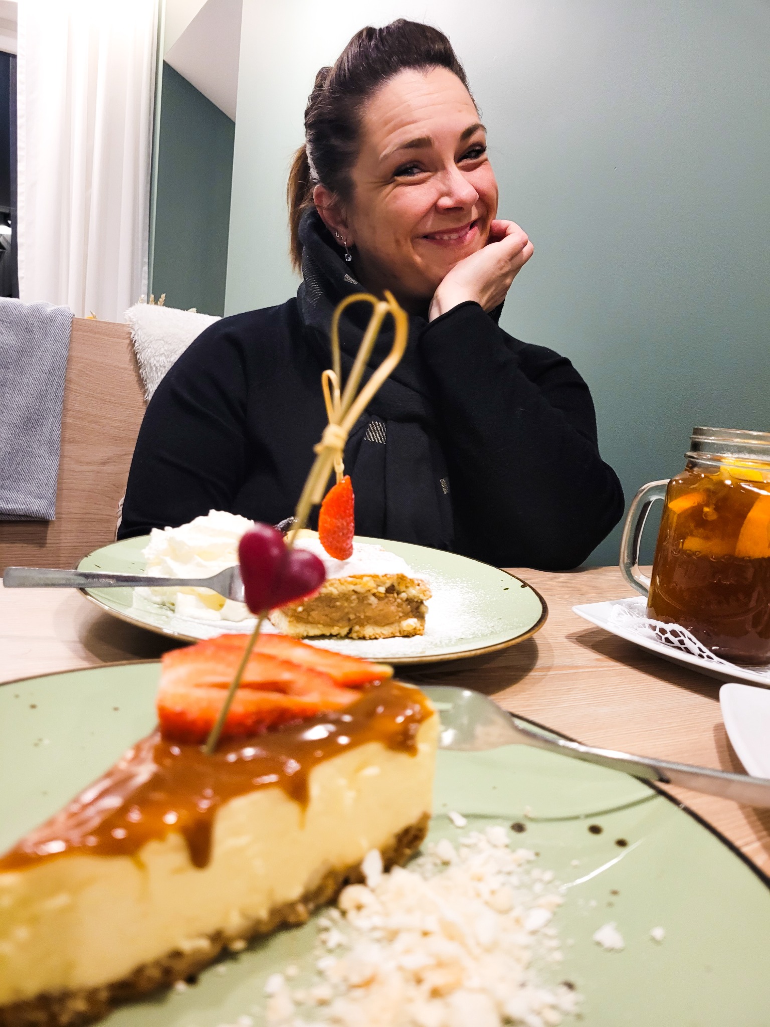 Uśmiechnięta kobieta siedzi przy stole z deserami: kawałek sernika z karmelem i truskawkami, ciasto z bitą śmietaną i napój w słoiku.