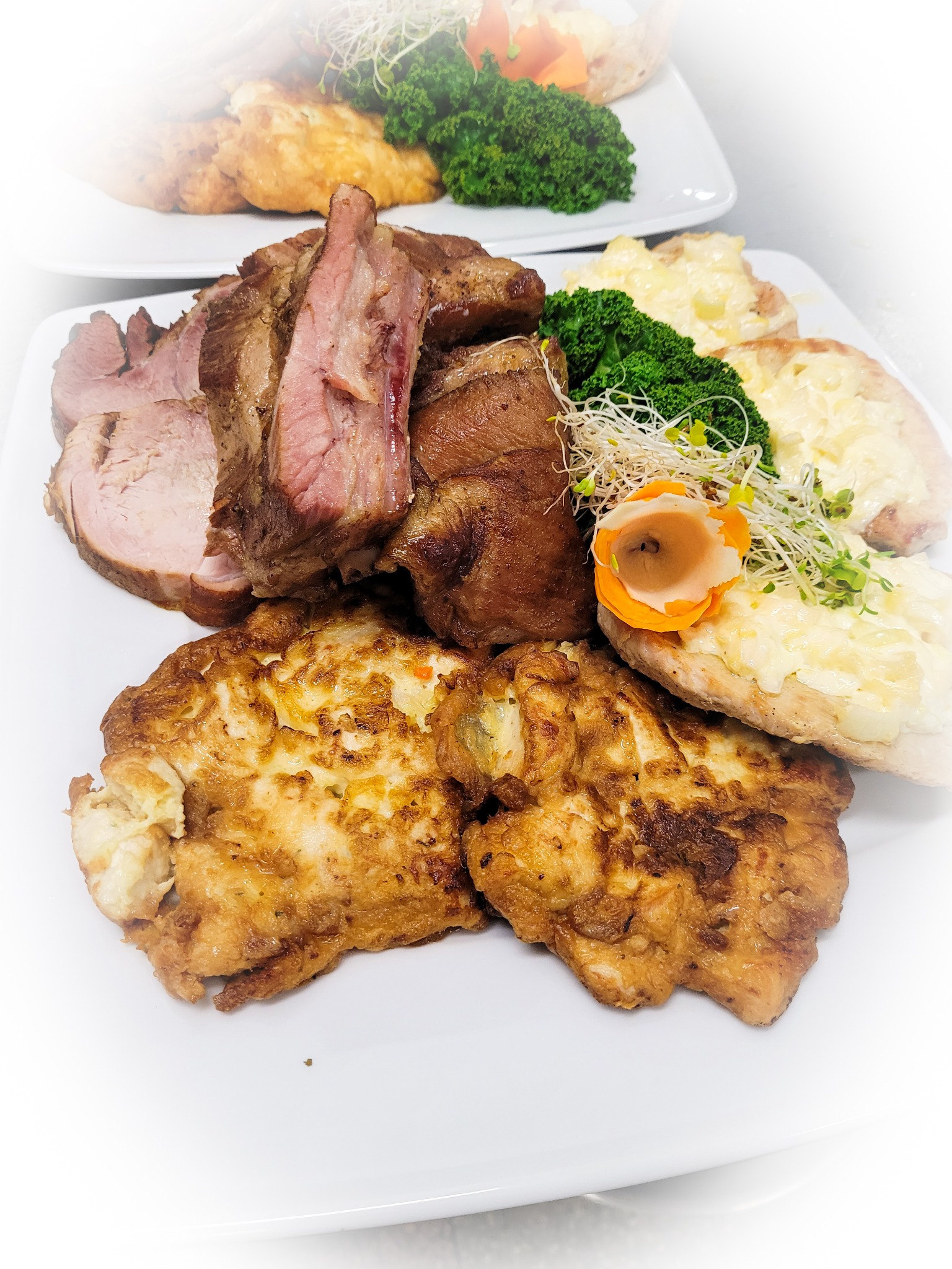 Dwa białe talerze z różnorodnymi daniami: pieczone mięso w plastrach, omlety, grzanki z serem, dekorowane brokułami, kiełkami i marchewką rzeźbioną w kwiat