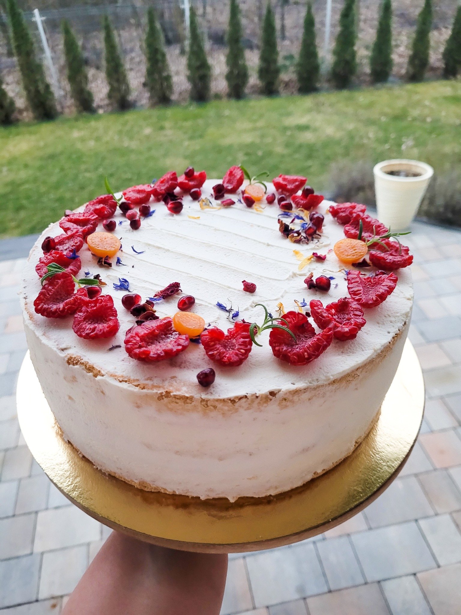 Tort z jasnym kremem, udekorowany malinami, granatem, physalis i suszonymi kwiatami, na złotym podkładzie, trzymany w dłoni, w tle zielony trawnik i tuje.