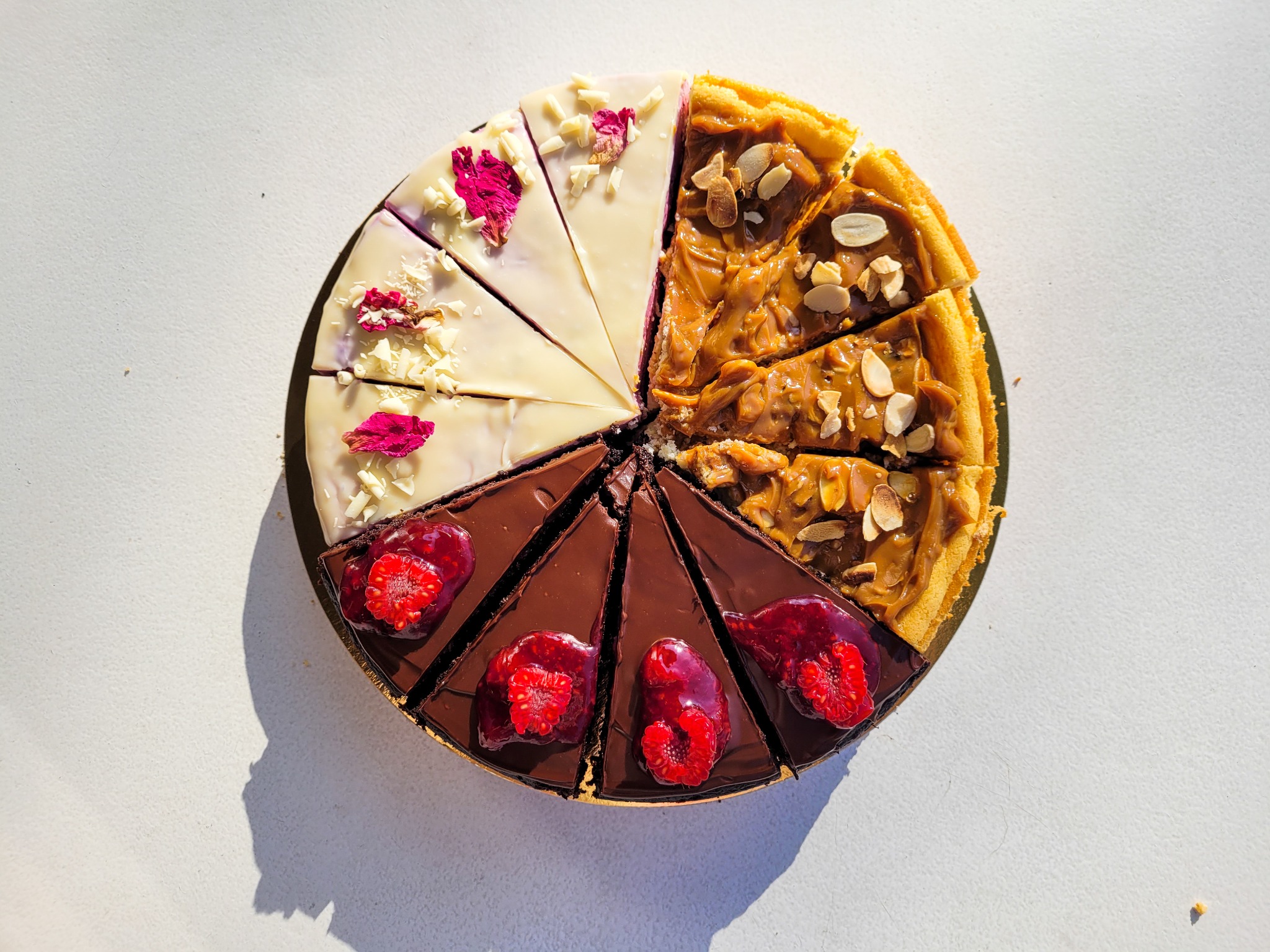 Kolaż ciast: czekoladowe z malinami w żelu, kajmakowe z migdałami i białe z płatkami róży i białą czekoladą, ułożonych na złotej tacy na białym tle w ostrym słońcu.