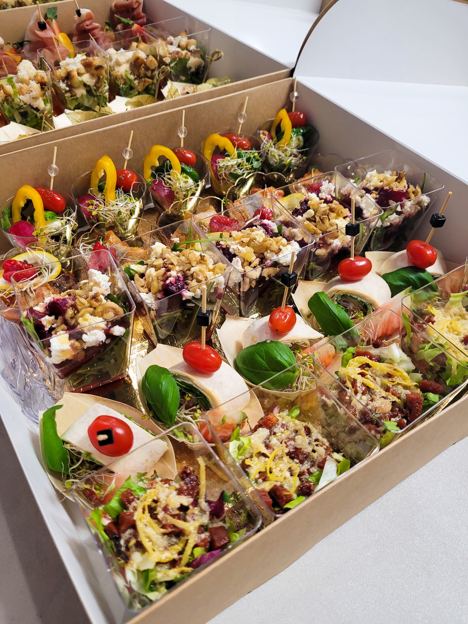 Zestaw różnorodnych przekąsek cateringowych w przezroczystych pojemnikach, umieszczonych w kartonowym pudełku, widoczne sałatki, roladki i koreczki, dekorowane świeżymi ziołami i warzywami.