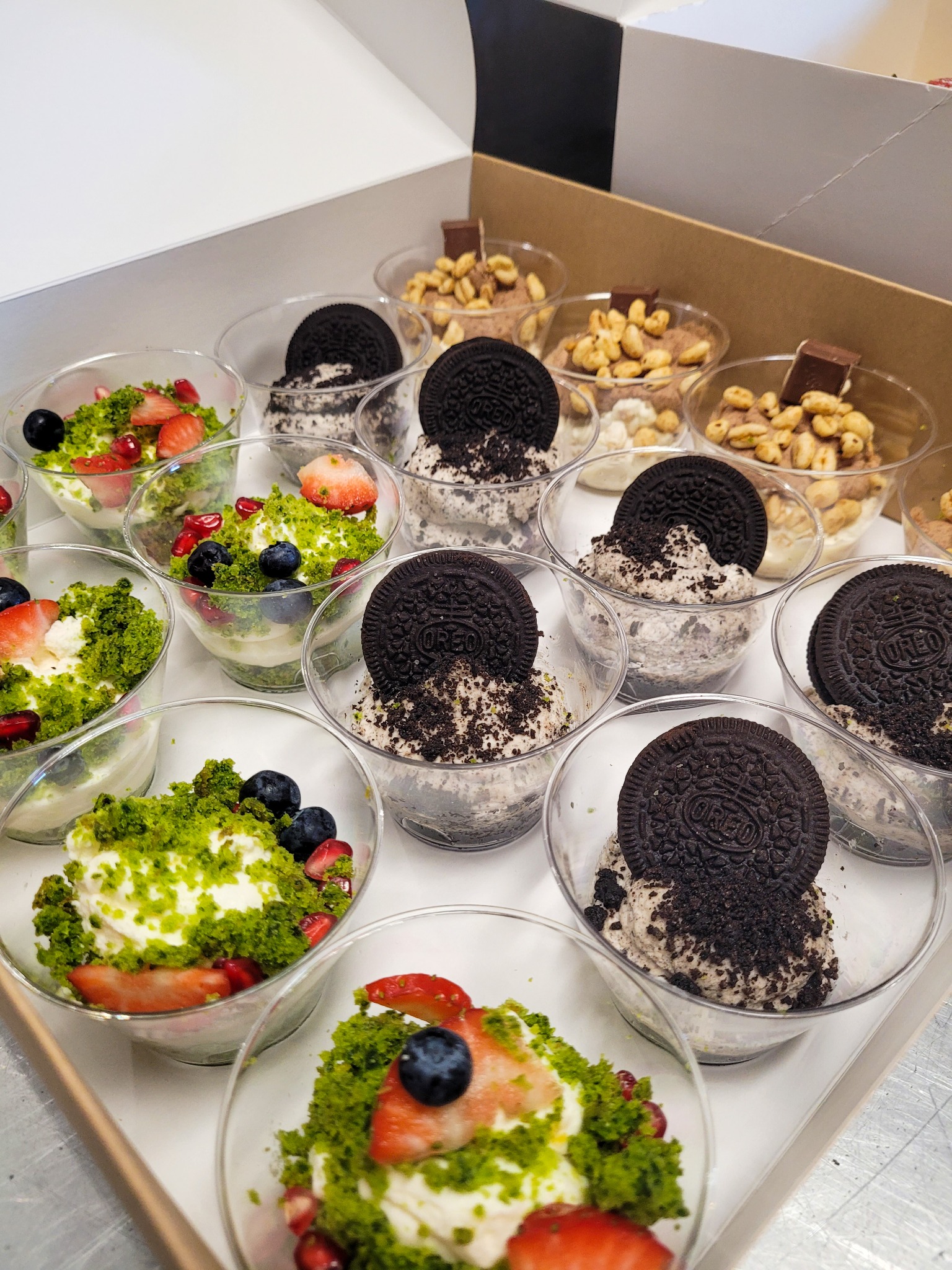 Deserki w plastikowych pucharkach w kartonowym pudełku: pucharki z kremem posypanym pistacjami i owocami (truskawki, borówki, granat) oraz pucharki z kremem z pokruszonymi ciastkami Oreo,...