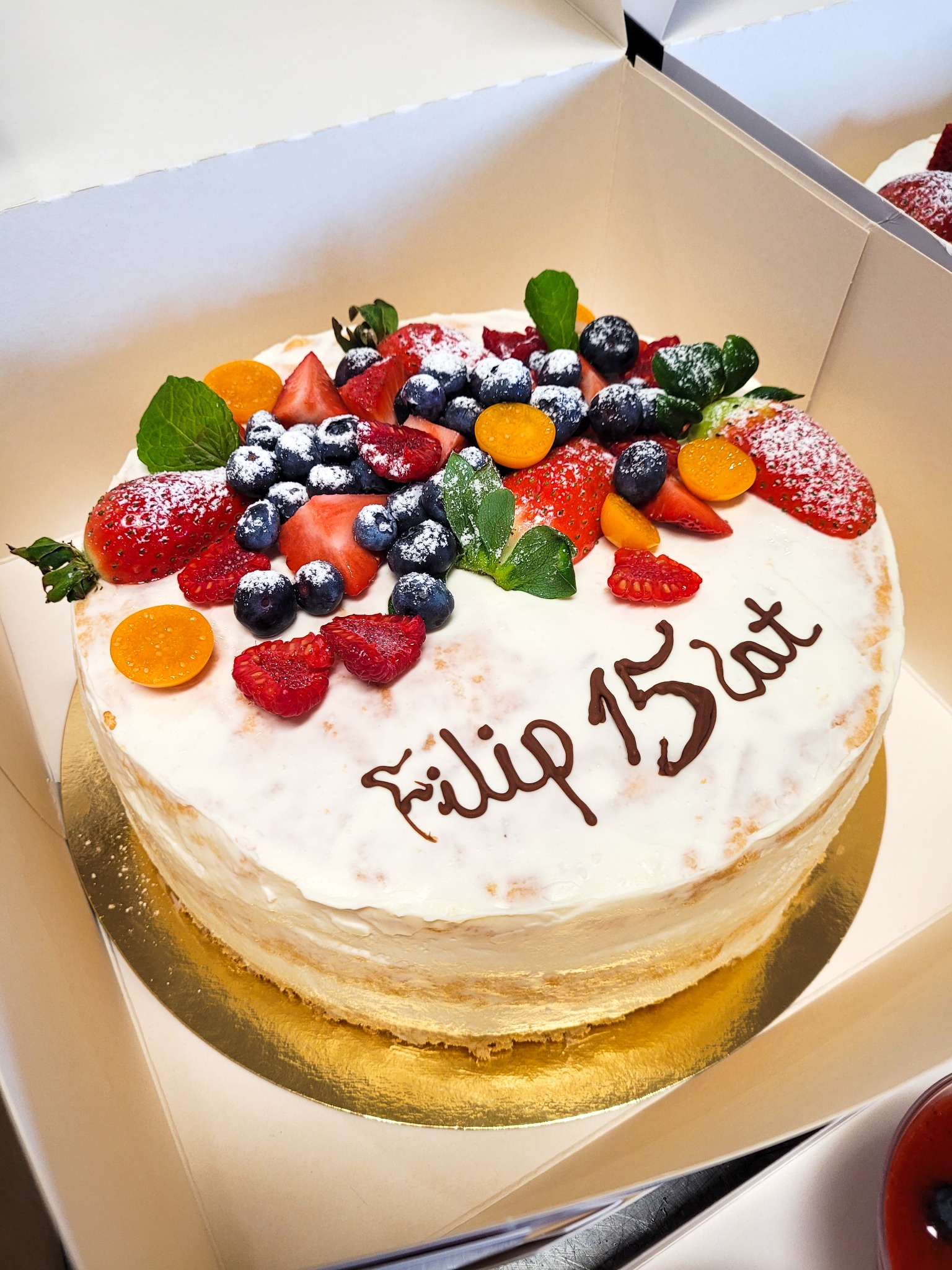 Tort urodzinowy z kremem, udekorowany świeżymi truskawkami, borówkami, malinami i miechunką, posypany cukrem pudrem, z napisem 'Filip 15 lat' wykonanym czekoladą, umieszczony w białym kartonie.