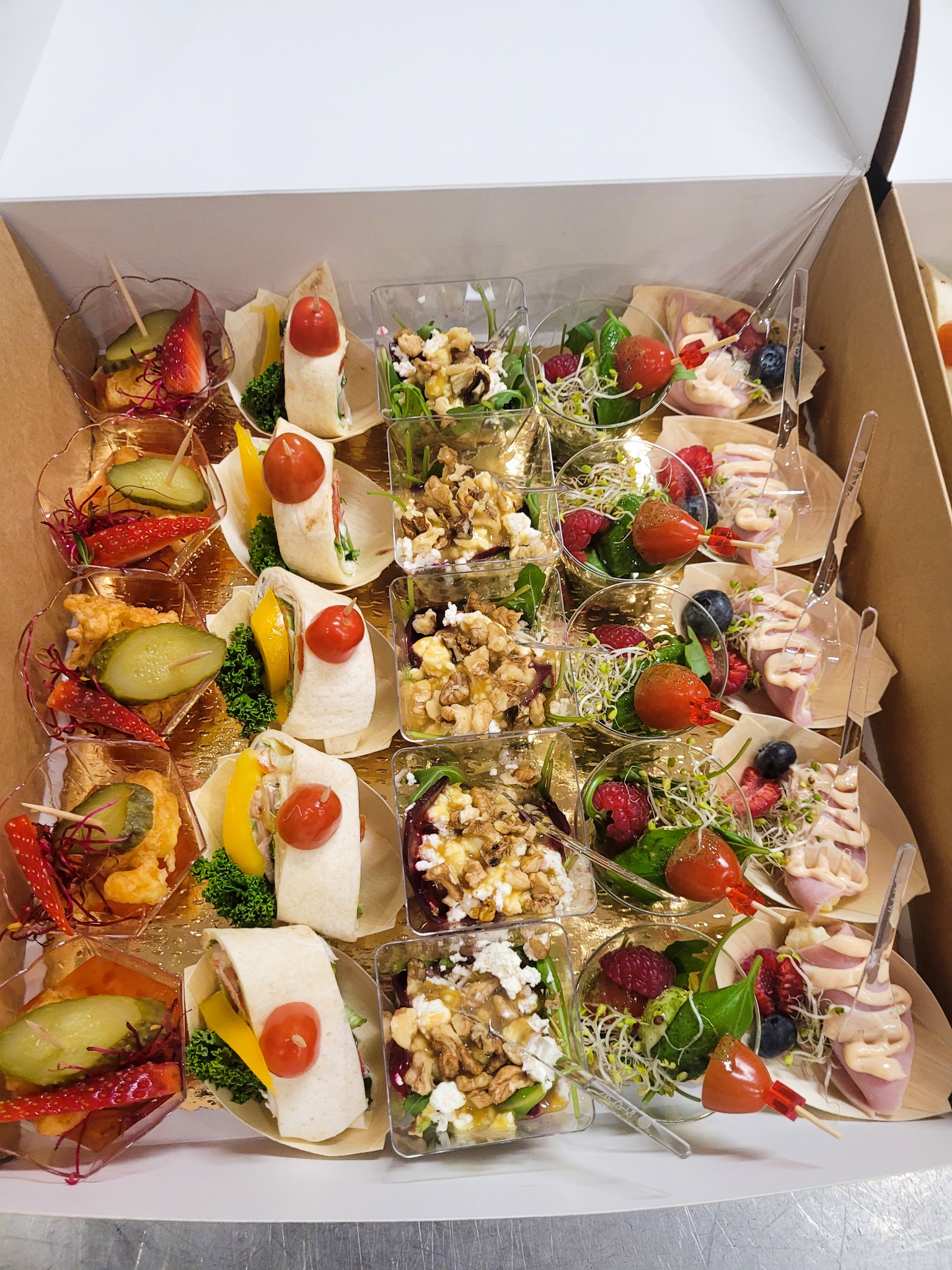 Zestaw różnorodnych przekąsek w pudełku cateringowym: mini tortille z warzywami, sałatki w szklanych naczyniach z orzechami i serem, koreczki z owocami i szynką, wszystko starannie ułożone i gotowe...