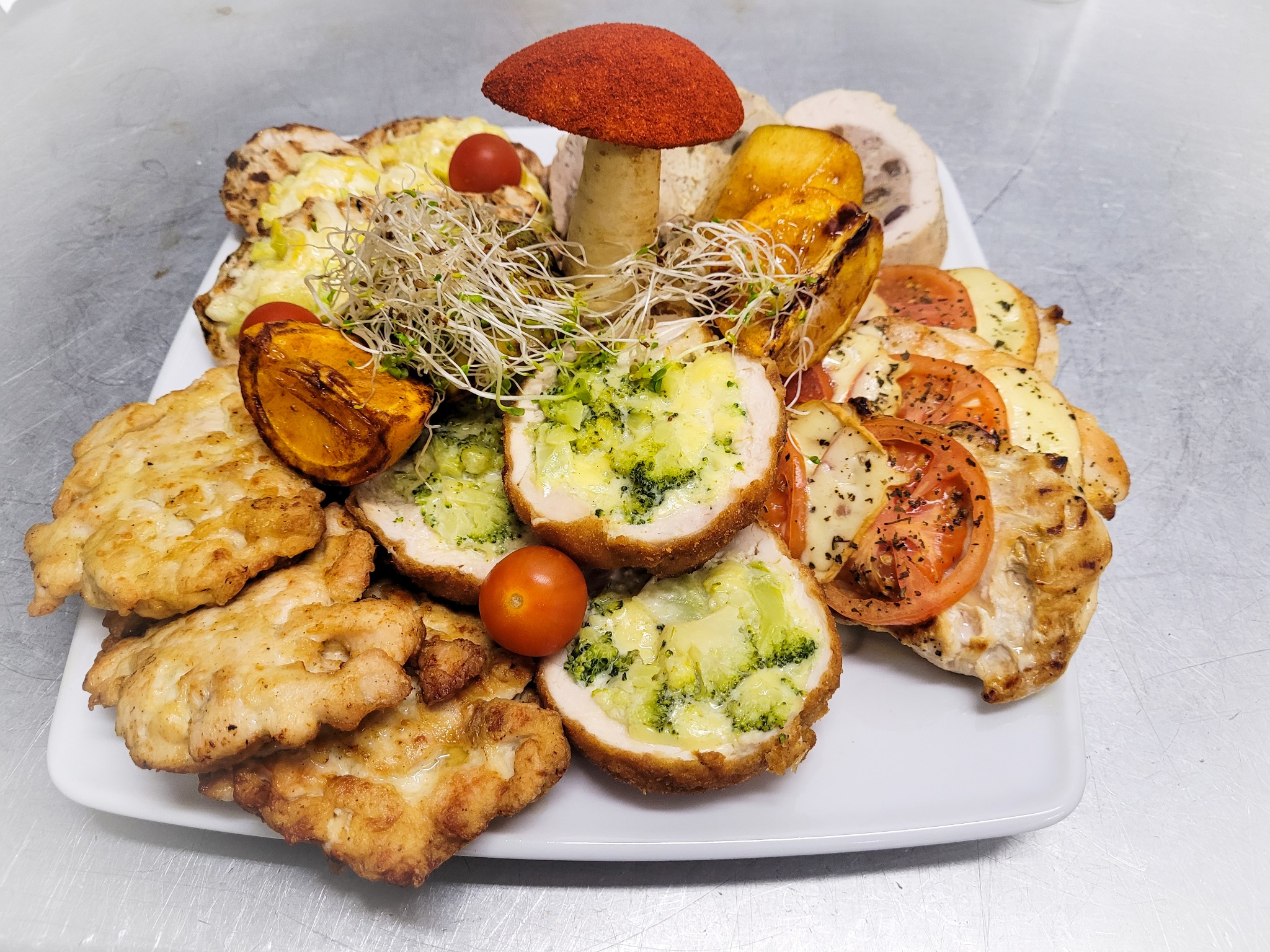 Dekoracyjna kompozycja potraw na białym talerzu: smażone kotlety, roladki z brokułami i serem, roladki z pomidorami i serem, pieczona dynia, grzyb z czerwonym kapeluszem i kiełki.