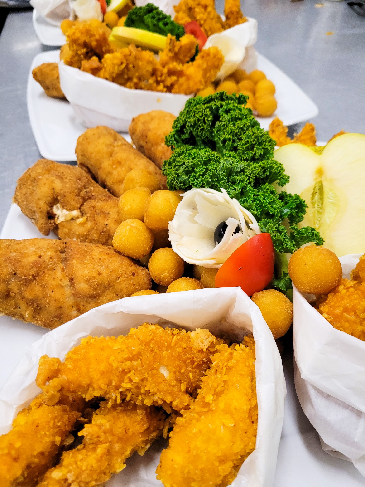 Kompozycja z panierowanych przekąsek, krokietów, smażonych kulek i dekoracji z warzyw na białych talerzach, ustawionych na metalowym blacie.