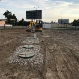 Żółta mini koparka Komatsu na placu budowy, w tle dwa zielone włazy studzienek kanalizacyjnych otoczone kamieniami, widoczne ślady opon na ziemi, w oddali tablice reklamowe i budynki.
