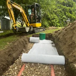 Żółta koparka Komatsu w wykopie z rurami drenażowymi, kamieniami i białą geowłókniną. Prace ziemne w ogrodzie, zielona trawa i drzewa w tle.