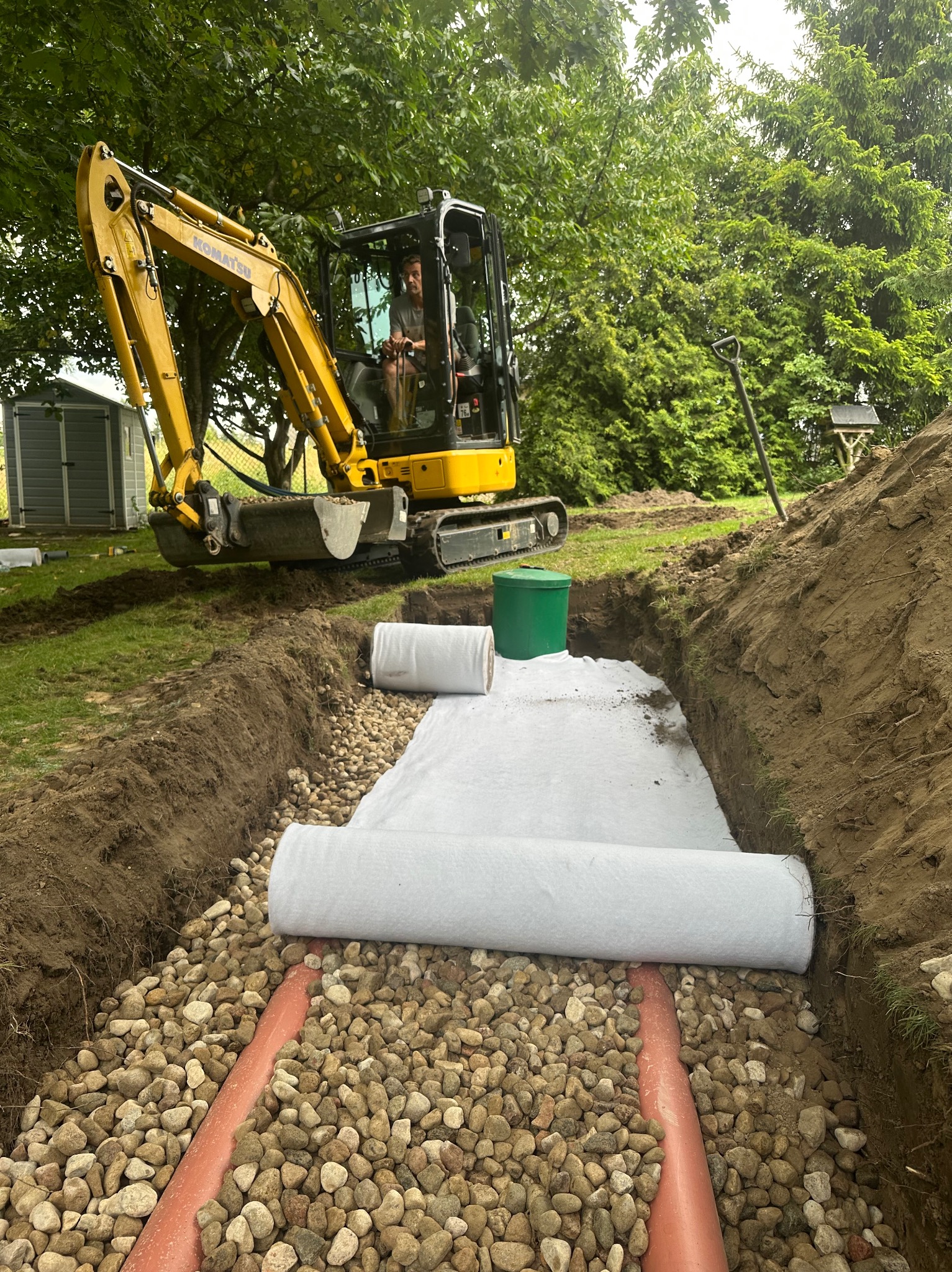 Żółta koparka Komatsu w wykopie z rurami drenażowymi, kamieniami i białą geowłókniną. Prace ziemne w ogrodzie, zielona trawa i drzewa w tle.