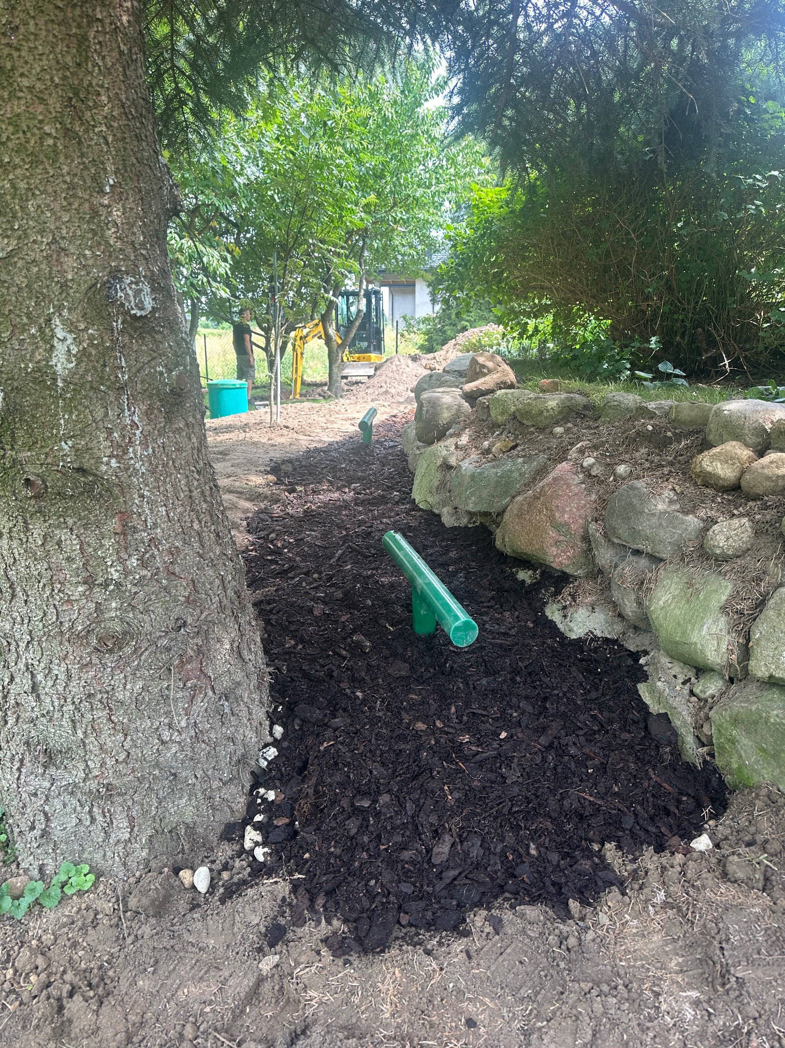 Wykop pod system nawadniania ogrodu, widoczne rury, ziemia i kora. W tle koparka i osoba. Realizacja z bliska, w słoneczny dzień.