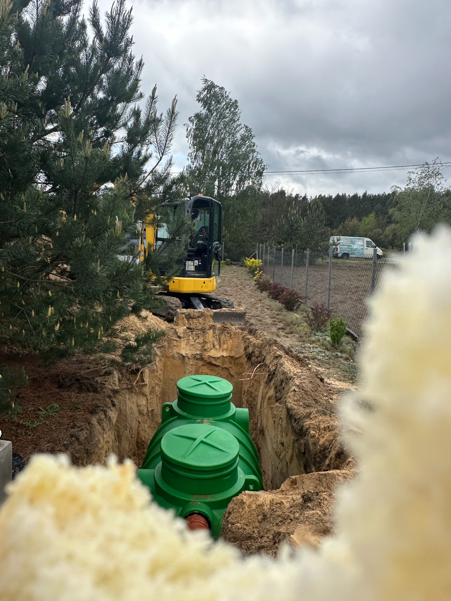 Zielony zbiornik w wykopie, widoczny zza przesłony. W tle minikoparka i ogrodzenie. Pochmurne niebo.