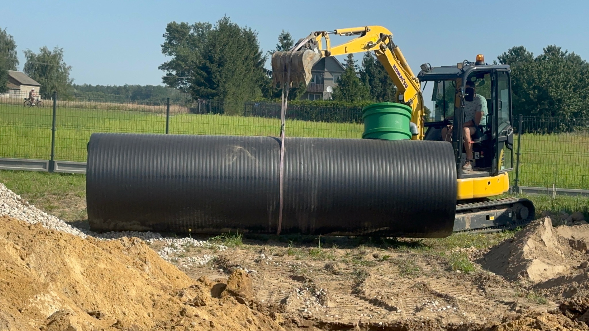Żółta koparka Komatsu umieszcza duży, czarny zbiornik na wodę deszczową w wykopie. Widoczny zielony element systemu, ziemia, kamienie i krajobraz w tle.