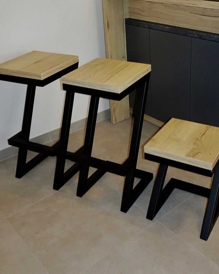 Trzy nowoczesne hokery z drewnianymi siedziskami i czarnymi, metalowymi nogami o geometrycznym kształcie, ustawione na jasnej podłodze w minimalistycznym wnętrzu z ciemnymi szafkami w tle.