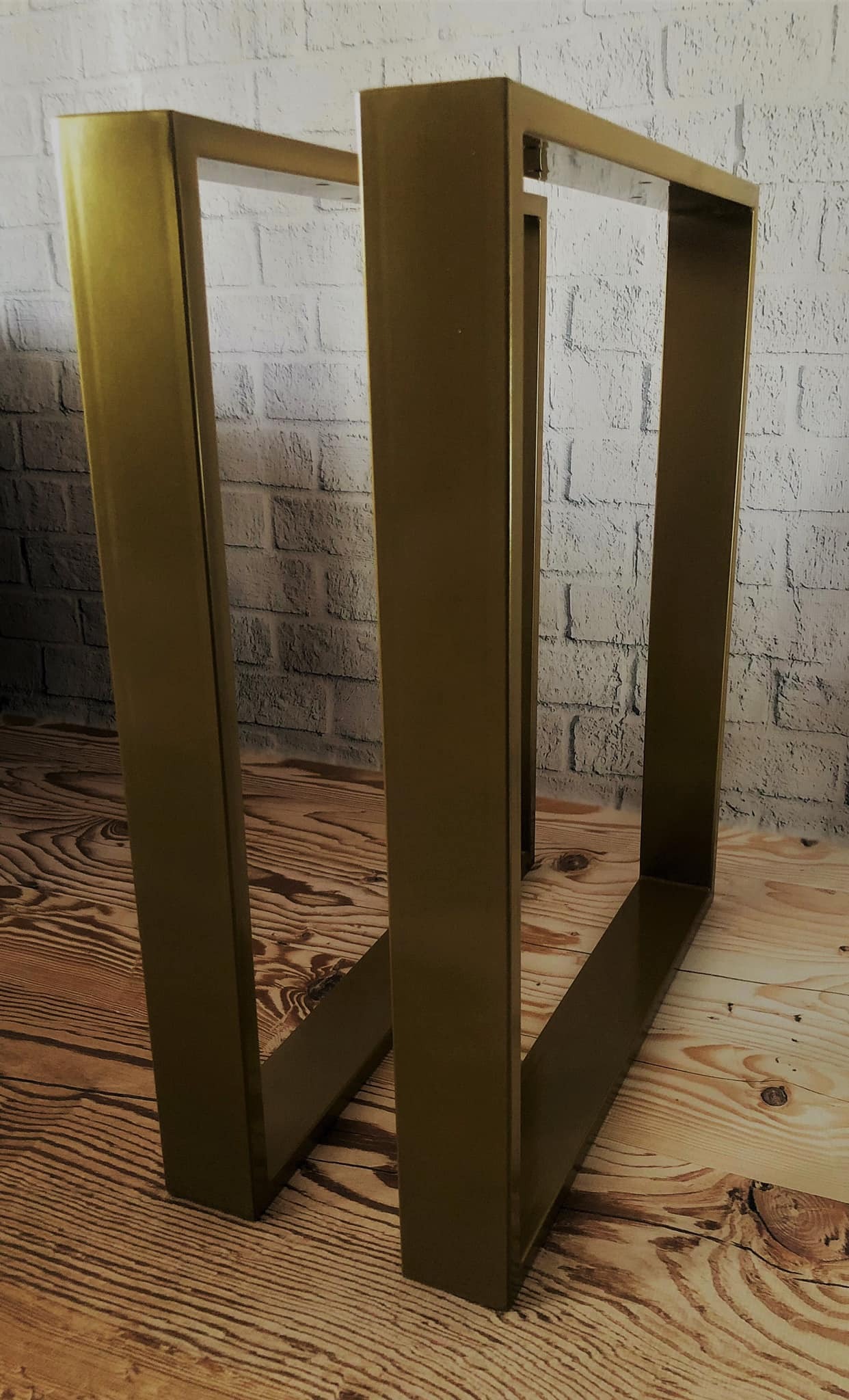Dwie metalowe, złote ramy w kształcie litery U, prawdopodobnie nogi do stołu, stoją na drewnianej podłodze na tle białej ściany z cegły.