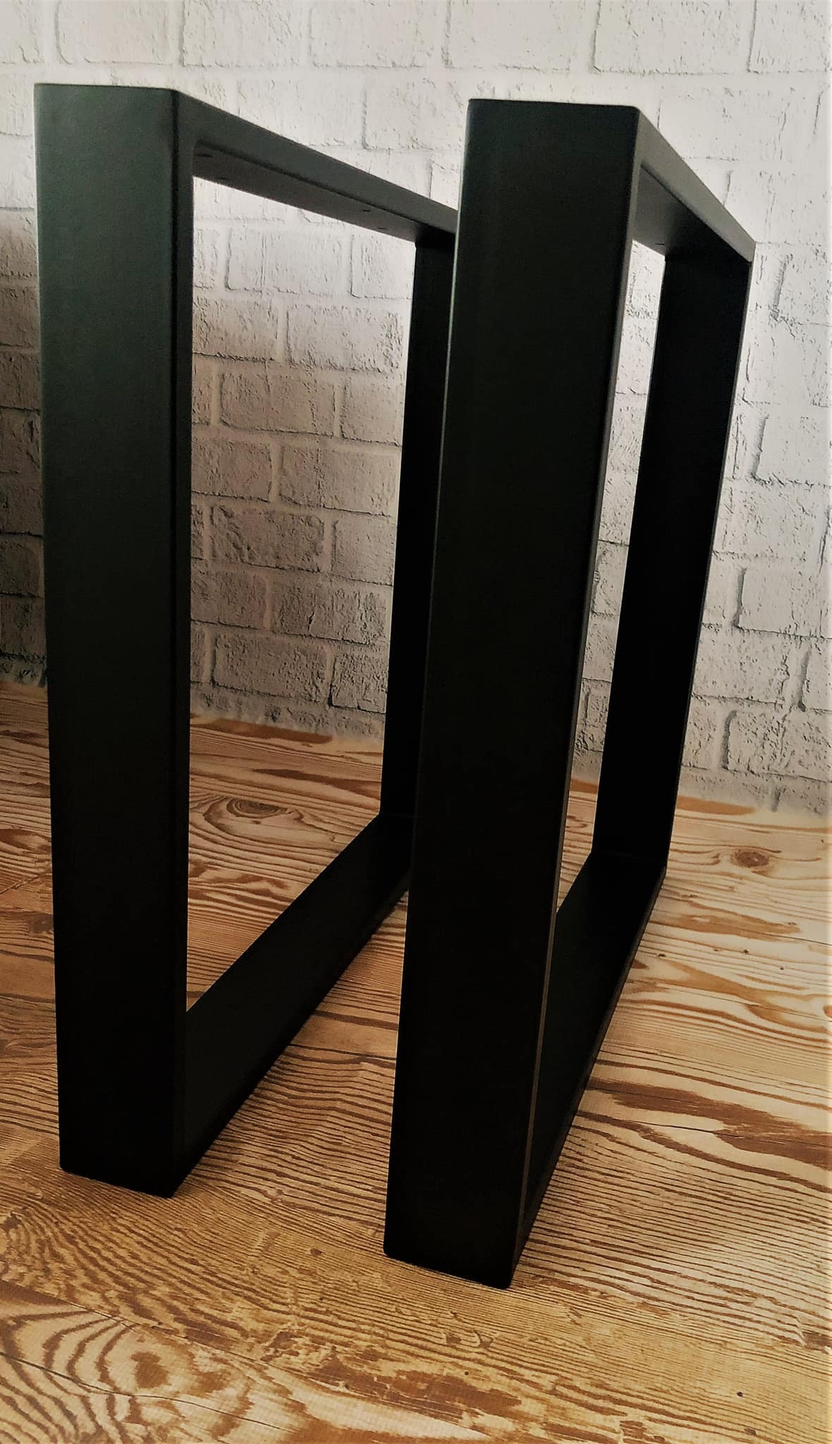Dwie czarne, metalowe podstawy stołu o prostokątnym przekroju, ustawione na drewnianej podłodze na tle ściany z białej cegły.