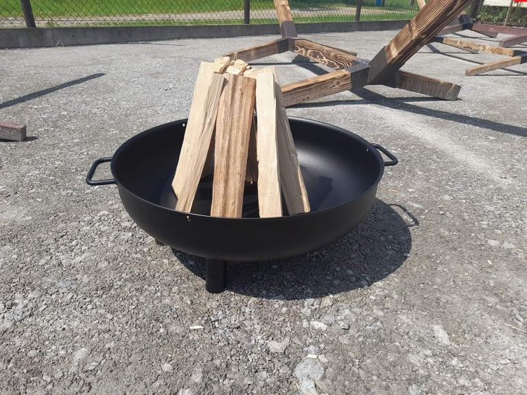 Czarny metalowy palenisko ogrodowe na trzech nóżkach, wypełnione ułożonym drewnem, na szarym żwirowym podłożu, w tle częściowo widoczna drewniana konstrukcja.