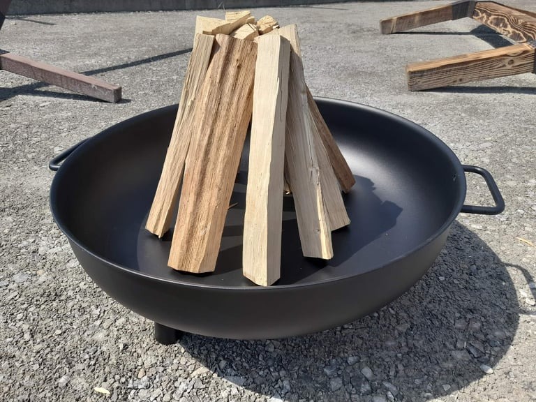 Czarna misa paleniskowa na trzech nóżkach, wypełniona ułożonymi kawałkami drewna na szarym, żwirowym podłożu. W tle fragmenty drewnianej konstrukcji.