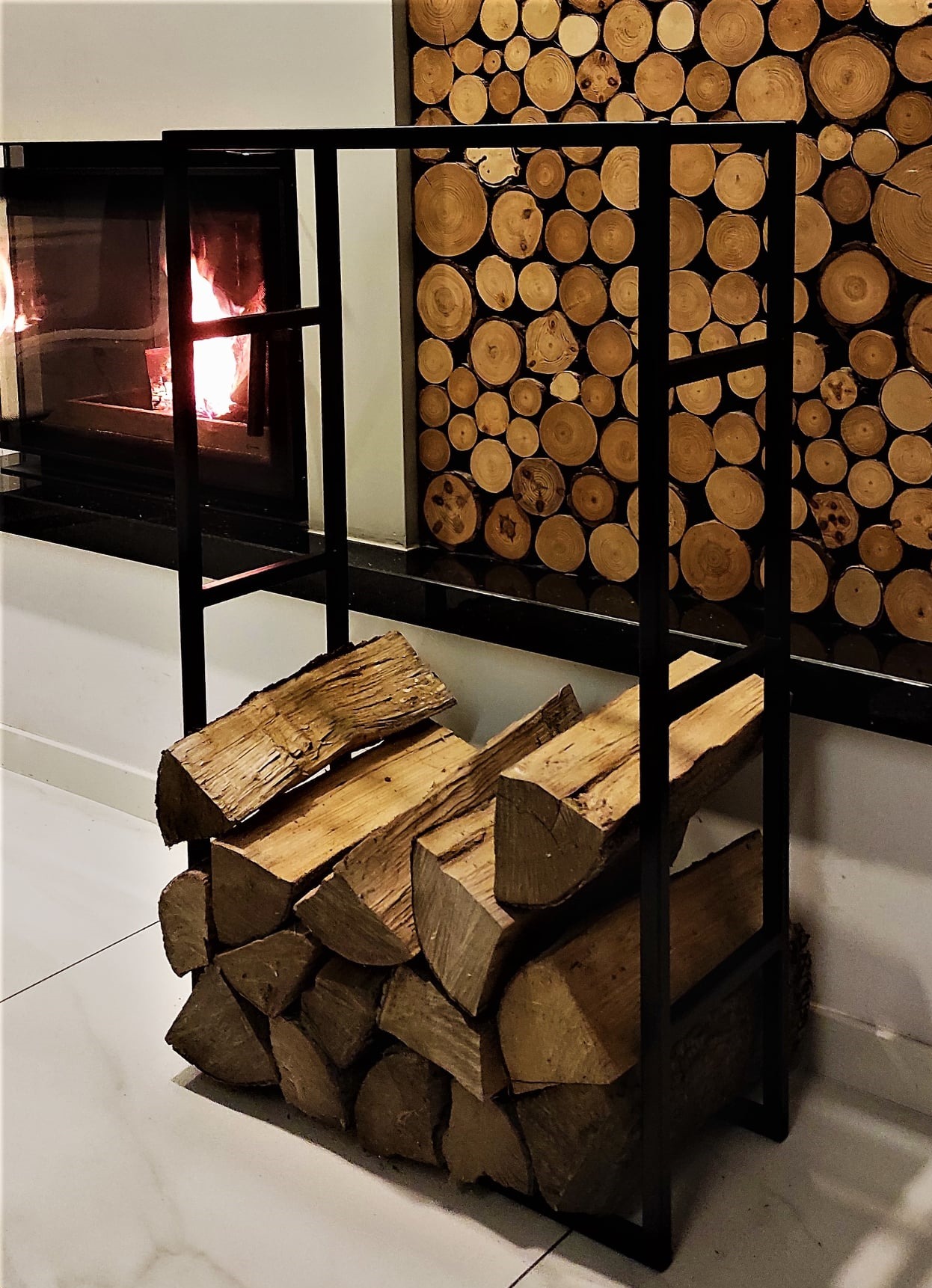 Metalowy stojak na drewno kominkowe, wypełniony pociętymi klockami drewna, na tle płonącego kominka i dekoracyjnej ściany z drewnianych krążków.