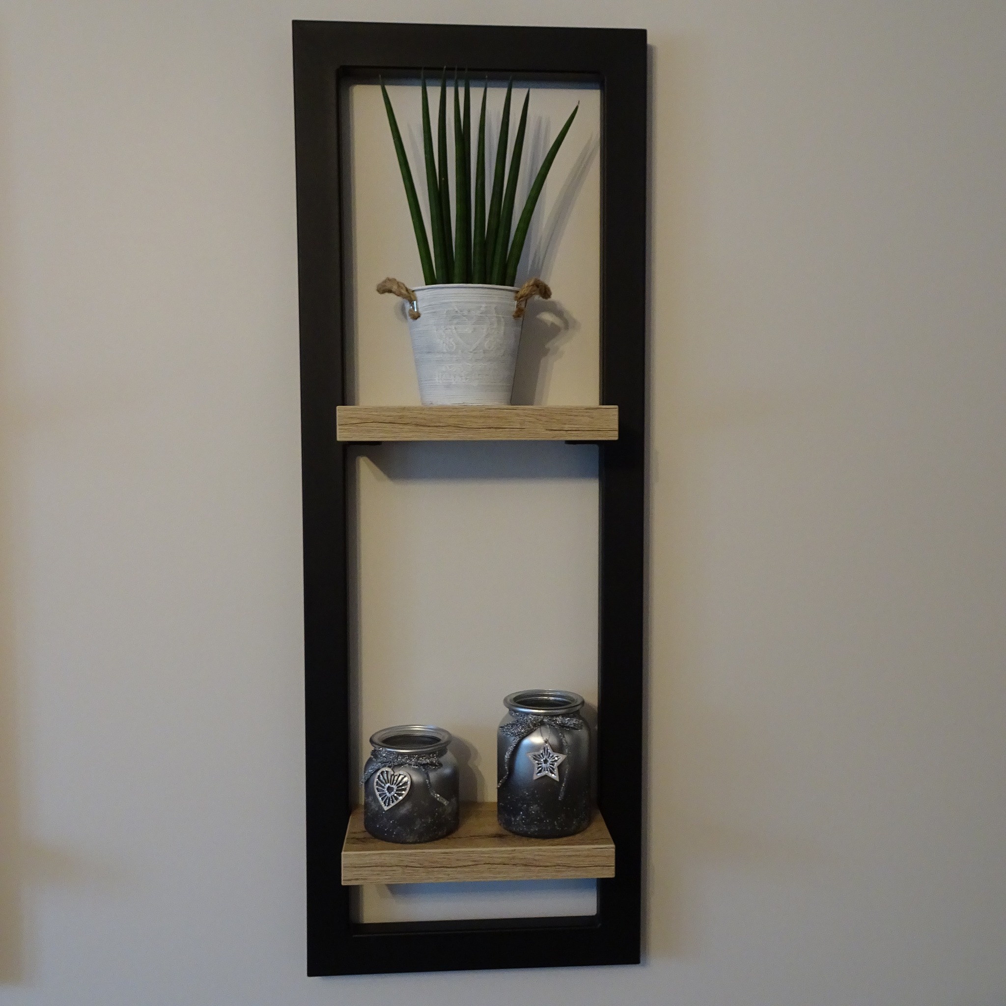 Półka ścienna w czarnej ramie z drewnianymi półkami, na których stoją dekoracyjne słoiki ze srebrnymi zawieszkami w kształcie serca i gwiazdy, a na górnej półce doniczka z zielonymi roślinami.
