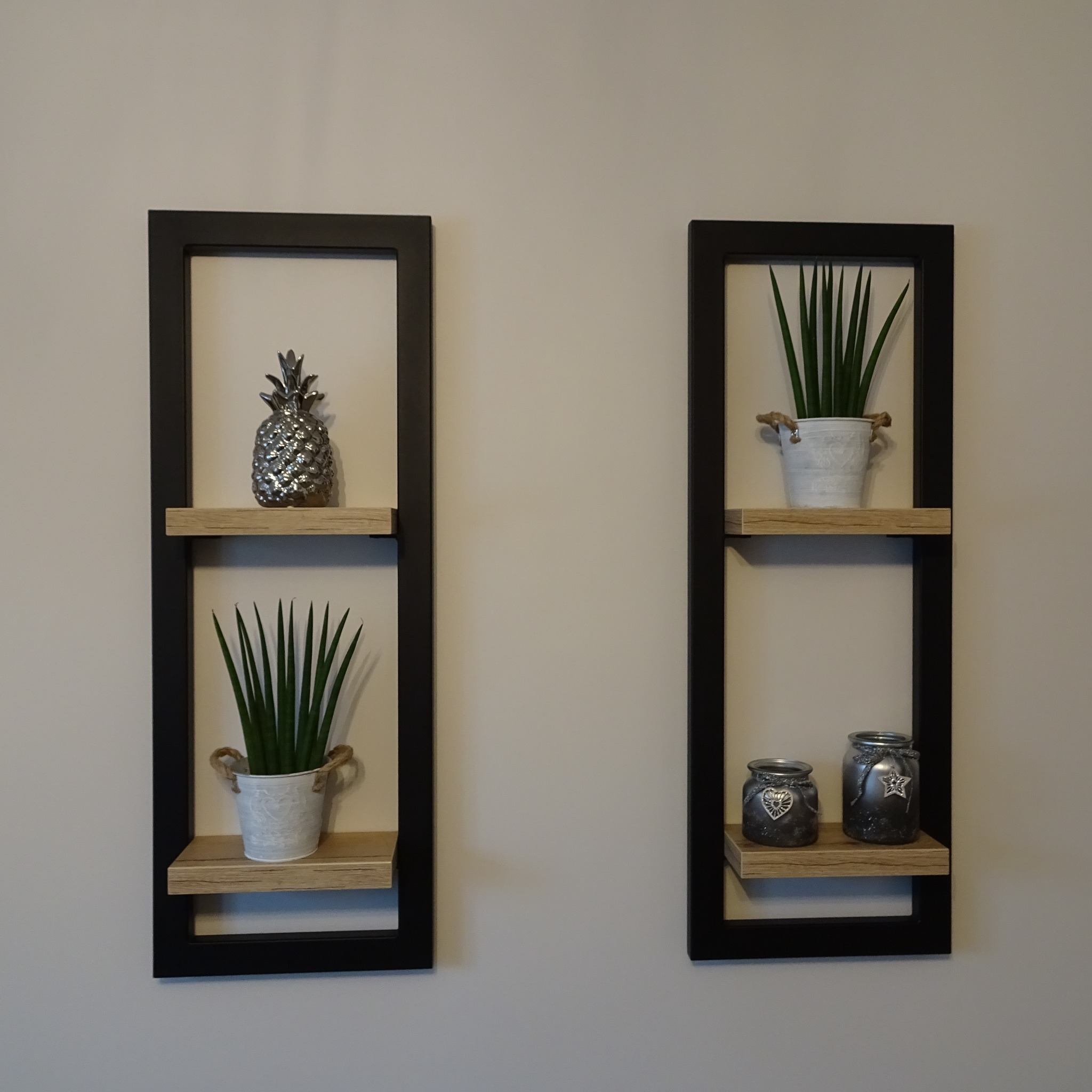 Dwie prostokątne, czarne półki wiszące z jasnego drewna, na ścianie w kolorze ecru. Na półkach dekoracje: srebrny ananas, dwie rośliny w białych doniczkach oraz dwa srebrne słoiczki.