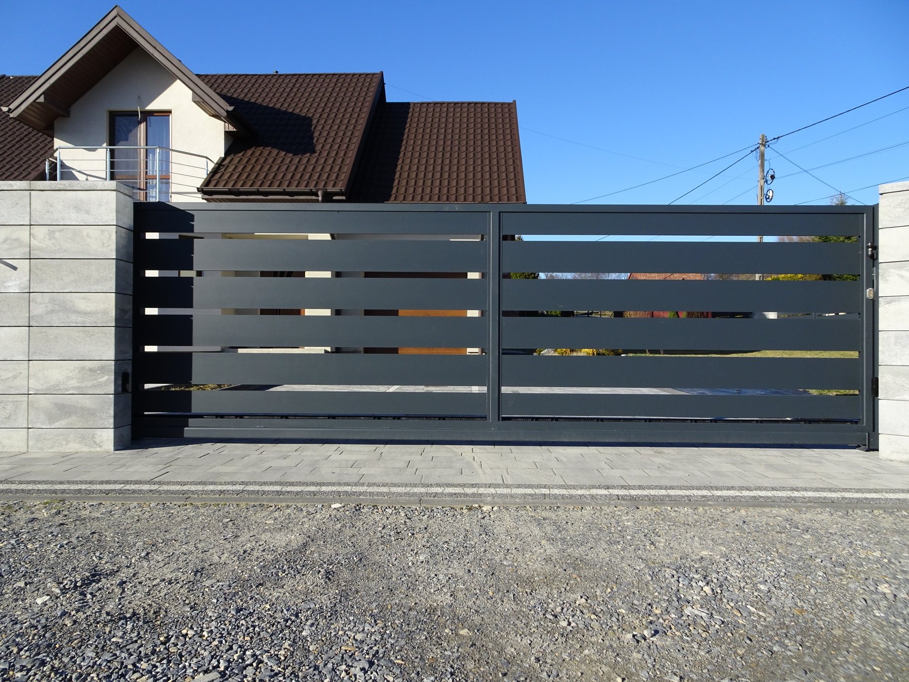 Nowoczesna, przesuwna brama wjazdowa w kolorze grafitowym z poziomymi szczeblami, zamontowana na betonowych słupkach z widocznym domem w tle.