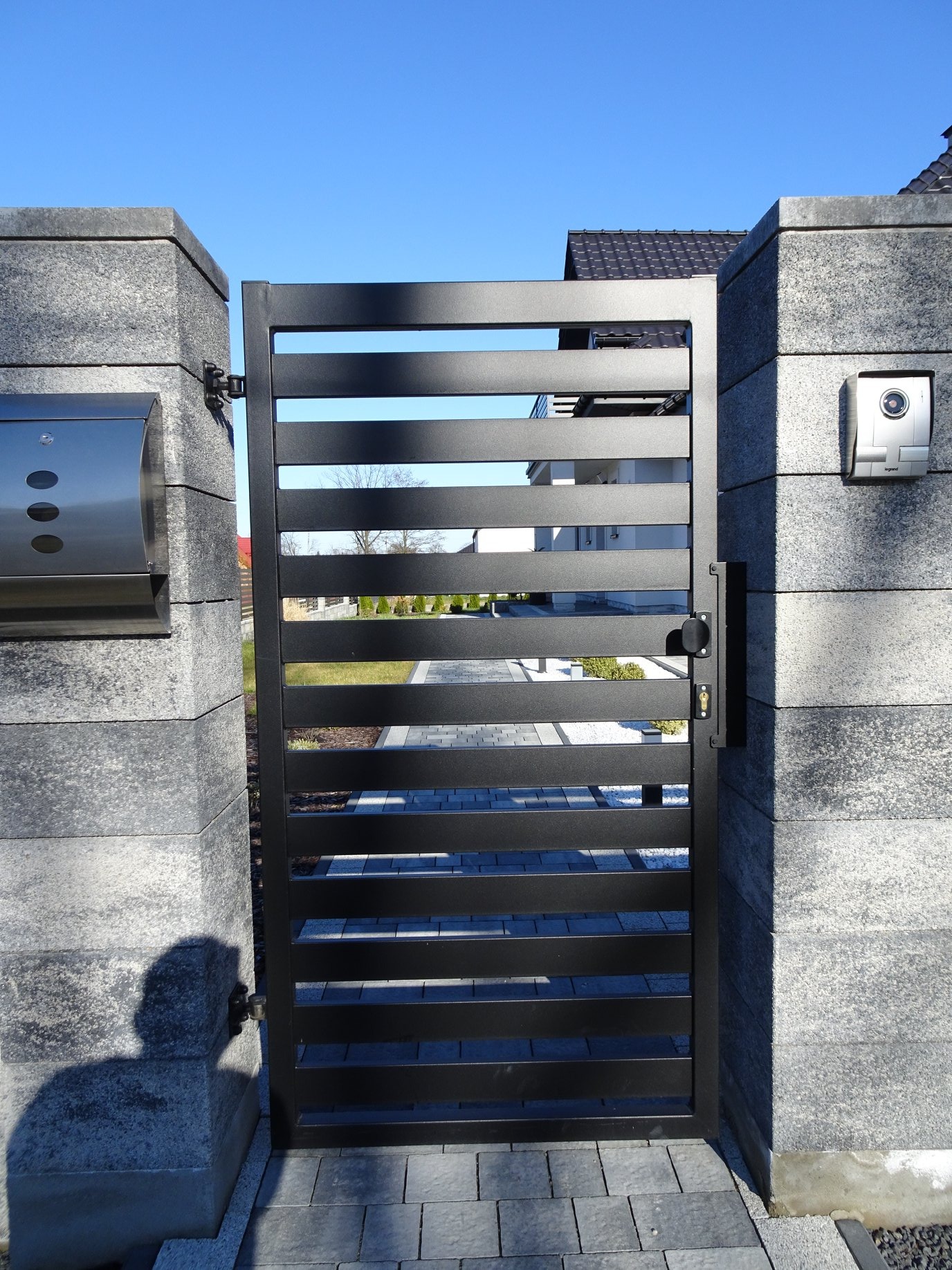 Nowoczesna, czarna brama z poziomych profili, zamontowana między kamiennymi słupkami z domofonem i skrzynką na listy, widok na posesję z kostką brukową i trawnikiem.