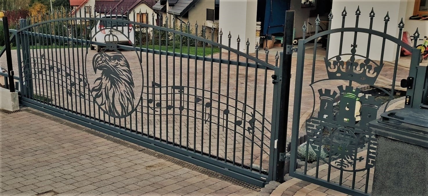 Otwarta, metalowa brama wjazdowa z dekoracyjnymi elementami wyciętymi laserowo: głowa orła wkomponowana w nuty oraz herb z koroną i zamkiem, na posesji z kostki brukowej.