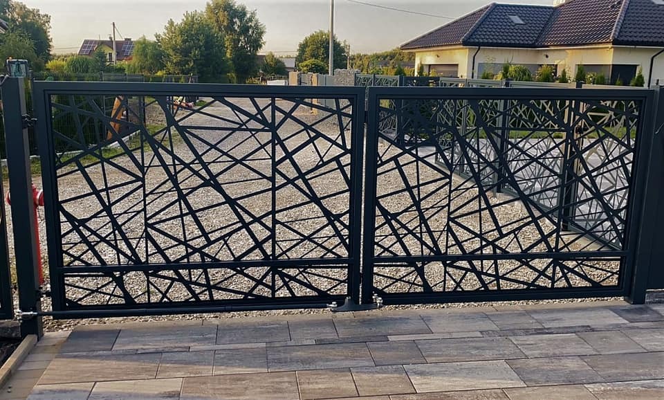 Nowoczesna brama wjazdowa dwuskrzydłowa, czarna, z geometrycznym ażurowym wzorem, na tle żwirowego podjazdu i domu z brązowym dachem.