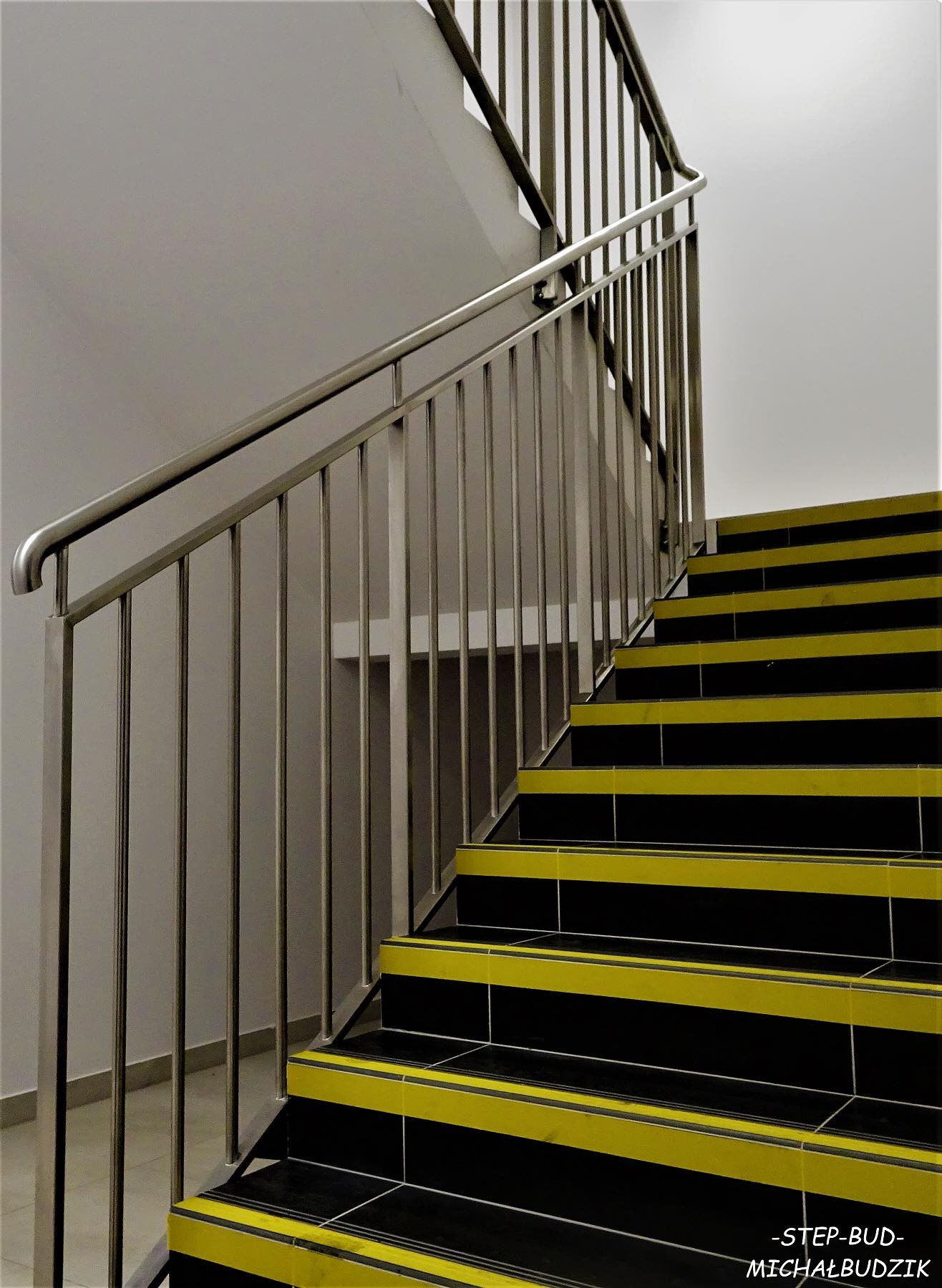 Klatka schodowa z metalową balustradą, stopnie wykończone czarnymi płytkami z żółtymi pasami antypoślizgowymi.