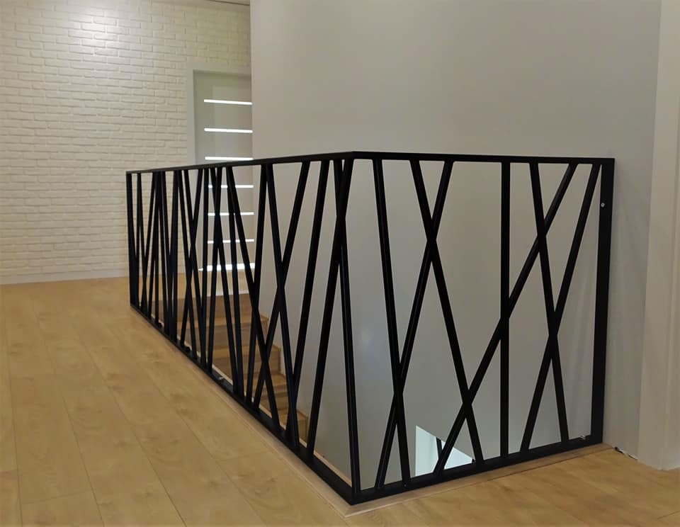 Czarna, metalowa barierka schodowa o geometrycznym wzorze krzyżujących się ukośnych prętów, zamontowana na jasnym parkiecie i białej ścianie z cegieł w tle.