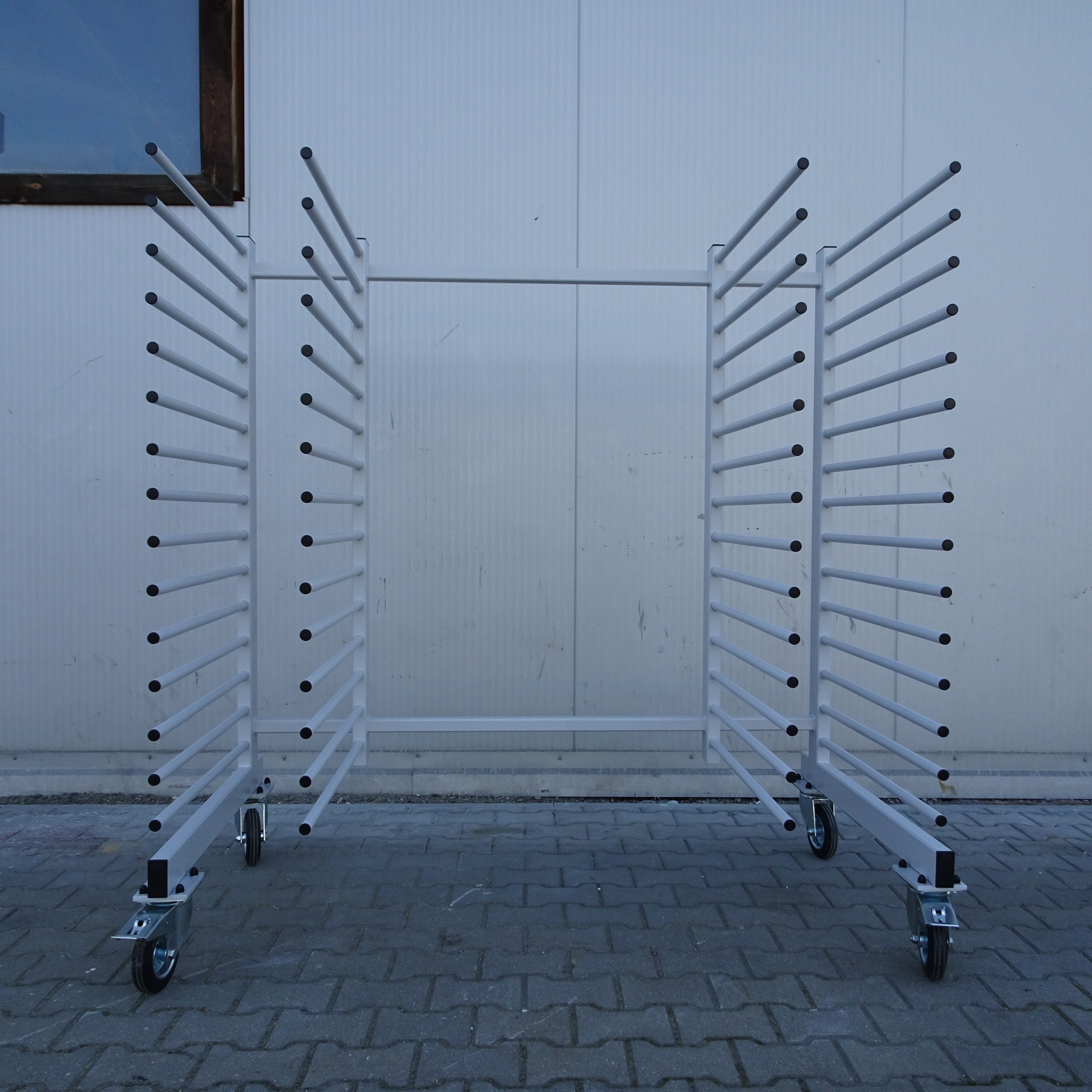 Mobilny, metalowy stojak na kółkach z poziomymi prętami, prawdopodobnie do suszenia lub przechowywania elementów po malowaniu, na tle białej ściany.