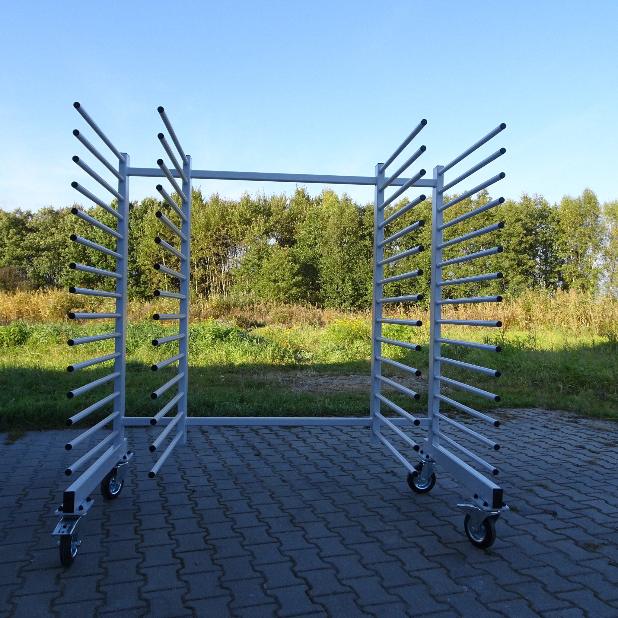 Metalowy, mobilny stojak z poziomymi prętami, prawdopodobnie do przechowywania lub transportu elementów, na tle zieleni i niebieskiego nieba.