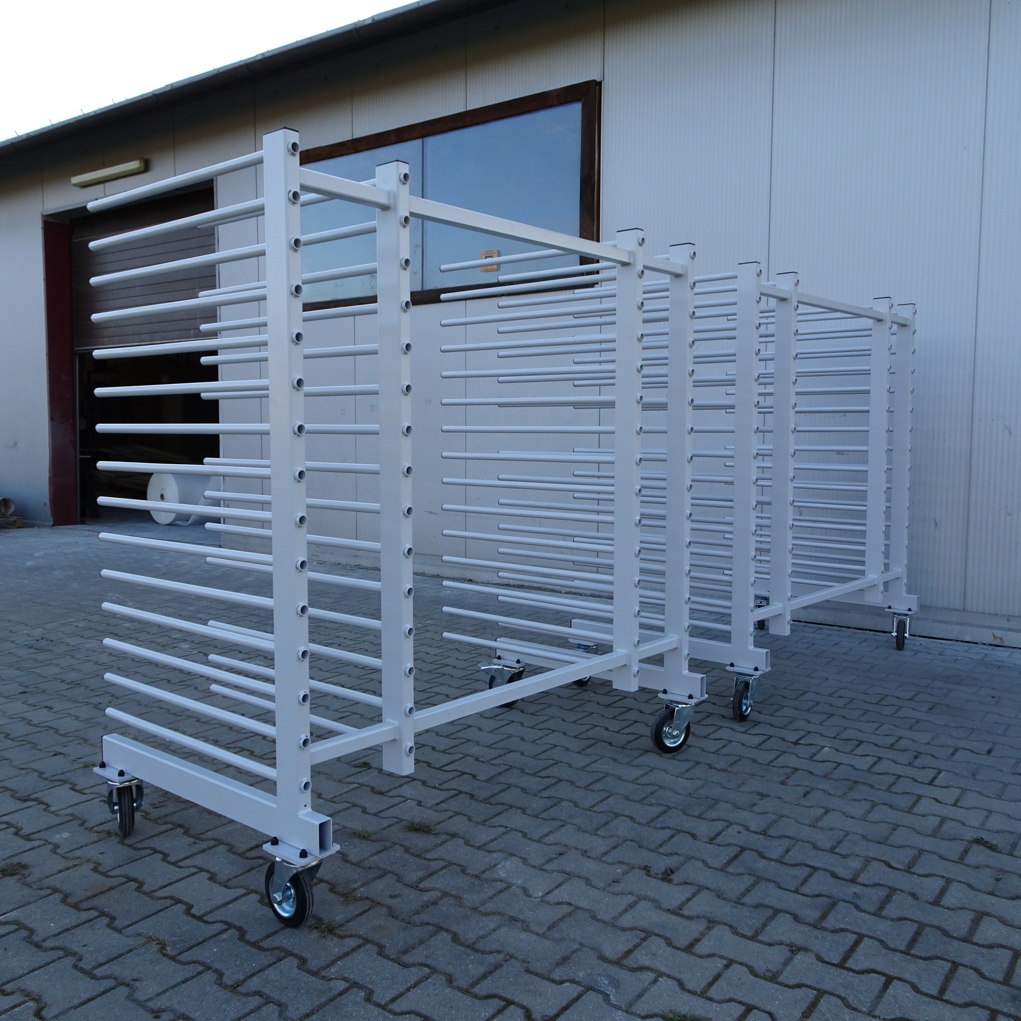 Wielopoziomowy, metalowy, mobilny regał warsztatowy na kółkach, z poziomymi prętami do suszenia lub przechowywania elementów, na tle budynku przemysłowego z oknem.