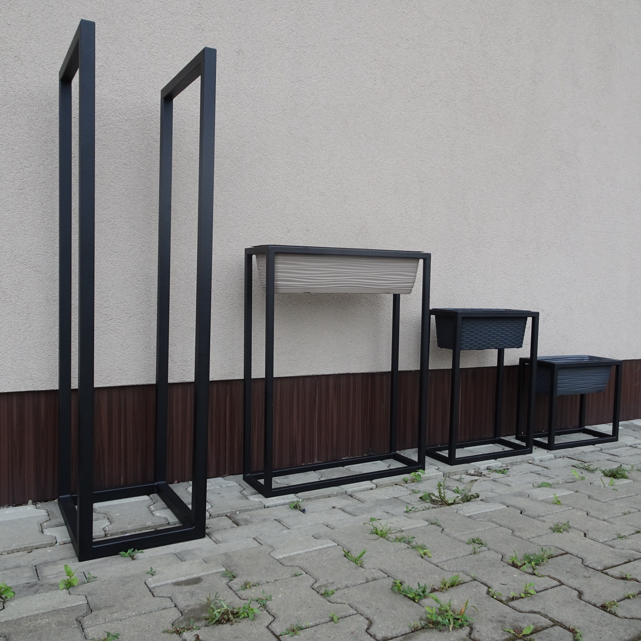 Zestaw trzech metalowych kwietników na podwyższeniu, o różnych wysokościach i rozmiarach donic, ustawionych na szarej kostce brukowej przed beżową ścianą.