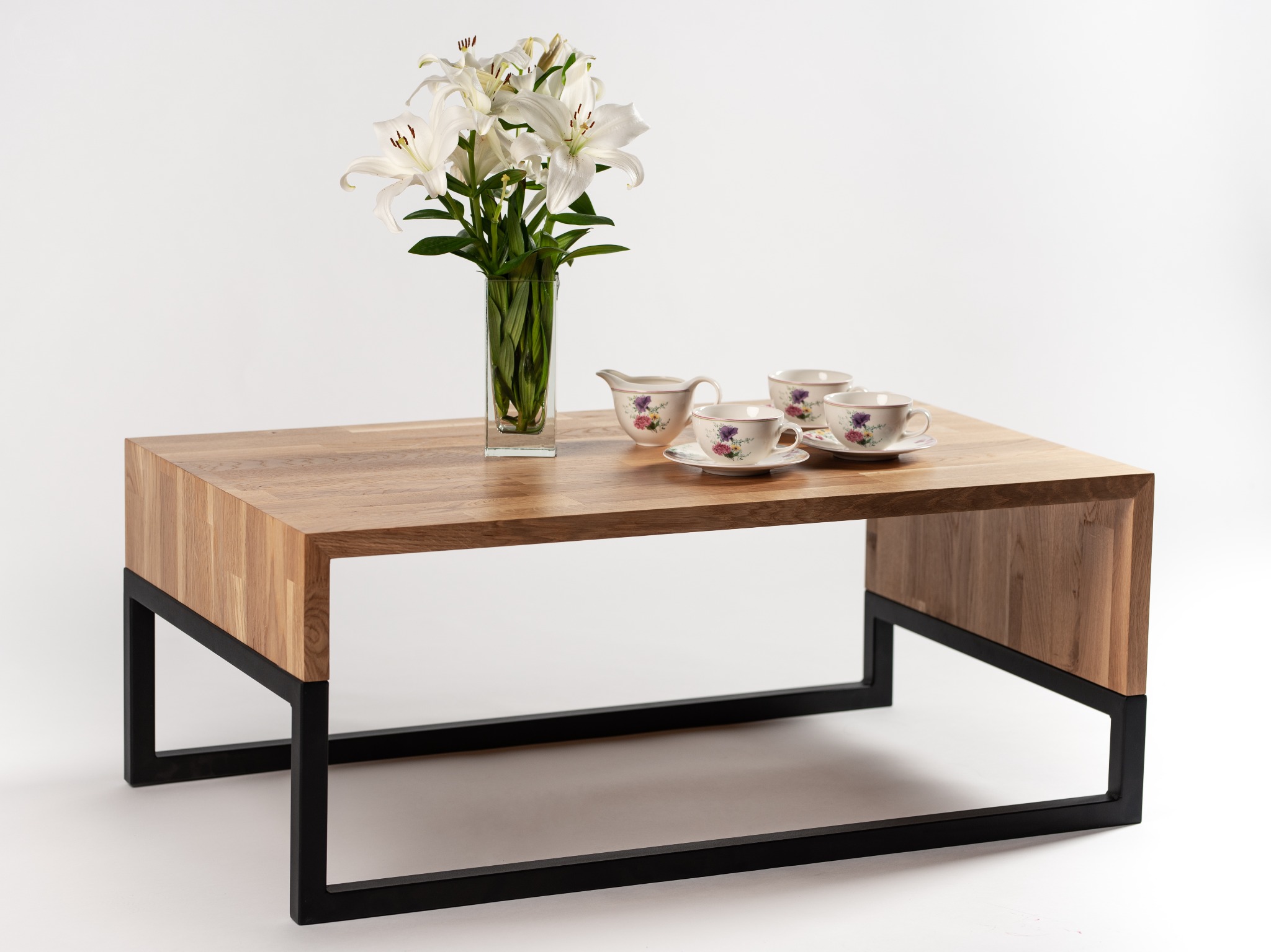 Stolik kawowy z blatem z litego drewna dębowego i czarną metalową podstawą, udekorowany wazonem z białymi liliami i zestawem porcelanowych filiżanek z kwiatowym motywem na białym tle.