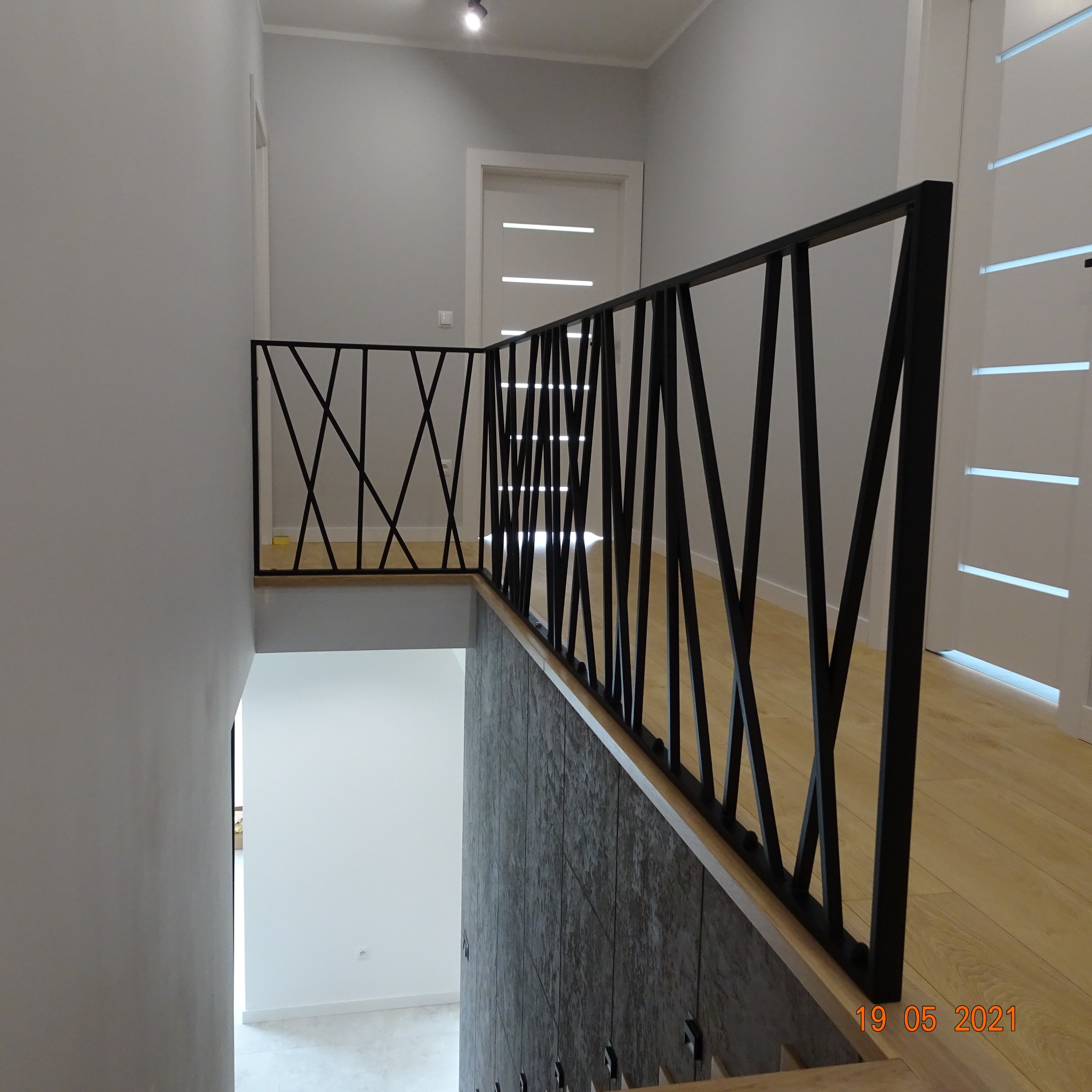 Czarna balustrada metalowa o geometrycznym wzorze w nowoczesnym wnętrzu z drewnianą podłogą i szarą ścianą, widok z góry na schody.