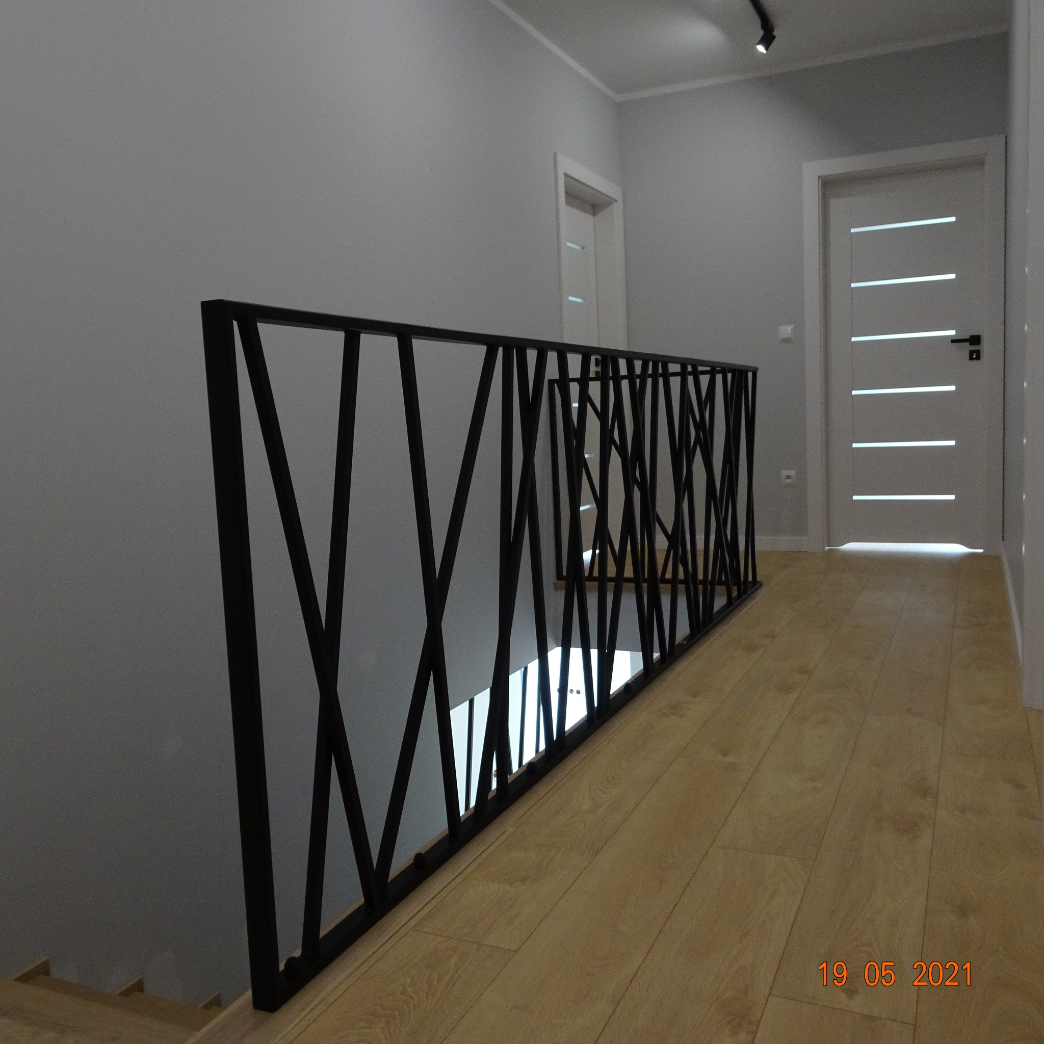 Nowoczesna czarna barierka schodowa z metalowych prętów krzyżujących się, na drewnianej podłodze w jasnym wnętrzu z białymi drzwiami.