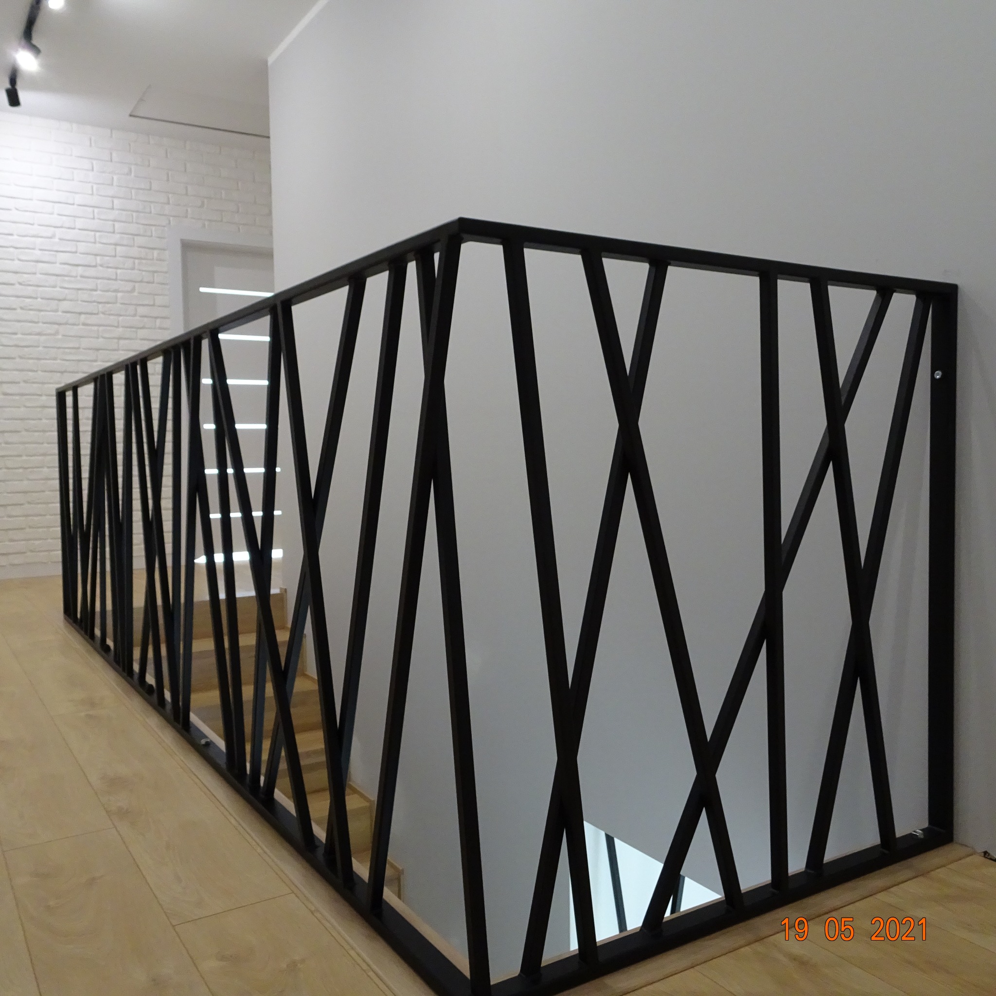 Nowoczesna balustrada schodowa z czarnych, ukośnych prętów, widok z boku na drewniane schody i parkiet, ściana z białej cegły w tle.