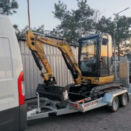 Żółta mini koparka CAT na przyczepie Brian James Trailers, zaparkowana obok białego furgonu na brukowanym placu. W tle widoczne drzewa, budynki i inne pojazdy.