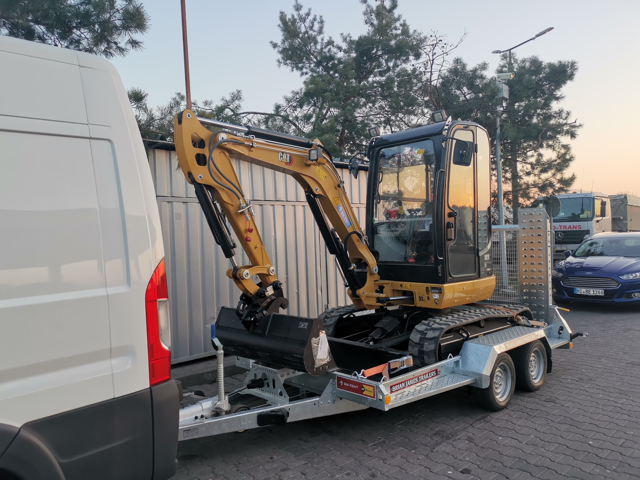 Żółta mini koparka CAT na przyczepie Brian James Trailers, zaparkowana obok białego furgonu na brukowanym placu. W tle widoczne drzewa, budynki i inne pojazdy.