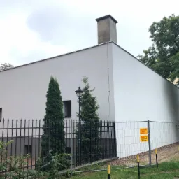Biała elewacja budynku z kominem, widoczna zza czarnego metalowego ogrodzenia i tymczasowego ogrodzenia budowlanego z żółtymi tablicami ostrzegawczymi, z drzewami w tle.