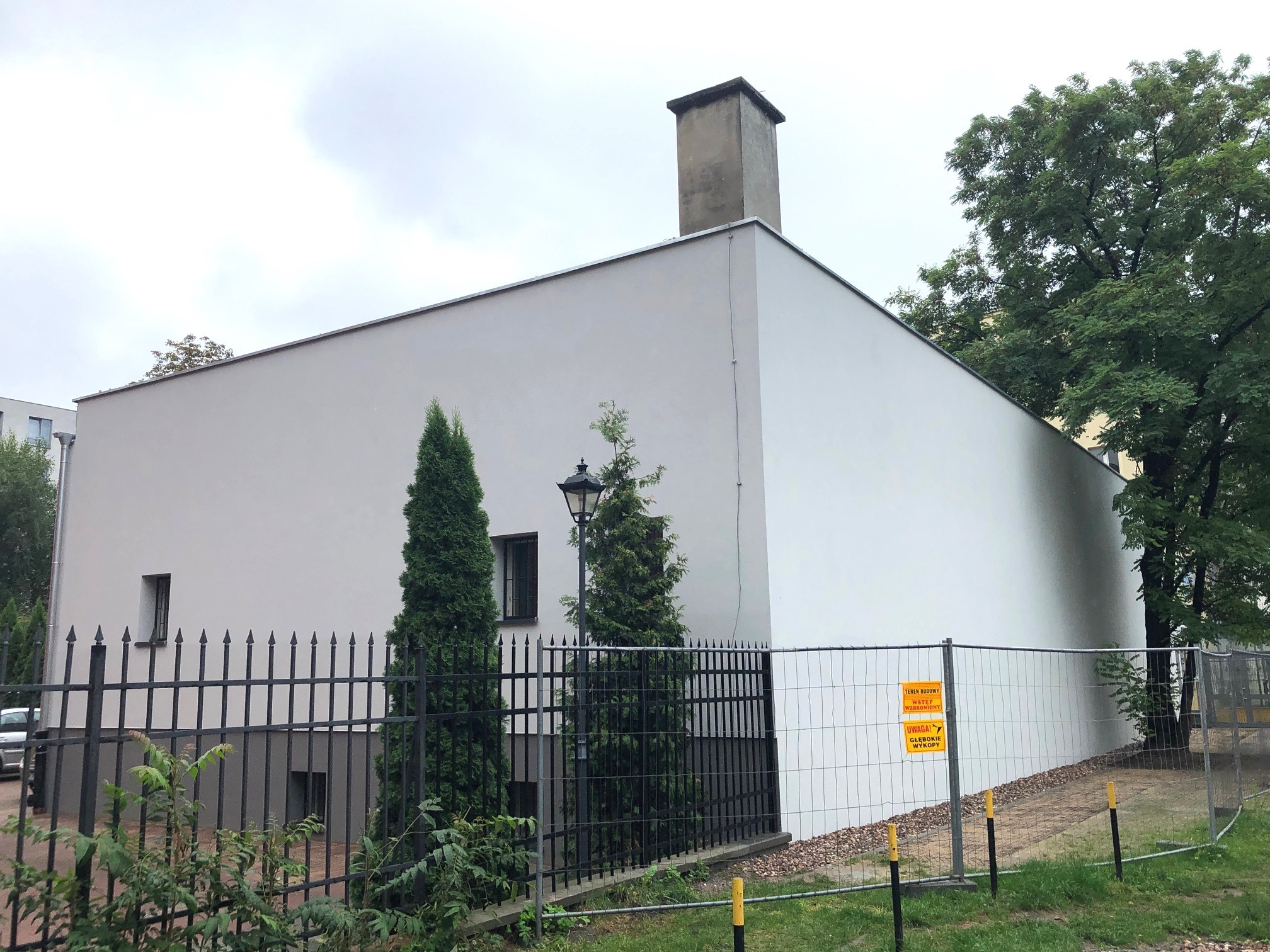 Biała elewacja budynku z kominem, widoczna zza czarnego metalowego ogrodzenia i tymczasowego ogrodzenia budowlanego z żółtymi tablicami ostrzegawczymi, z drzewami w tle.