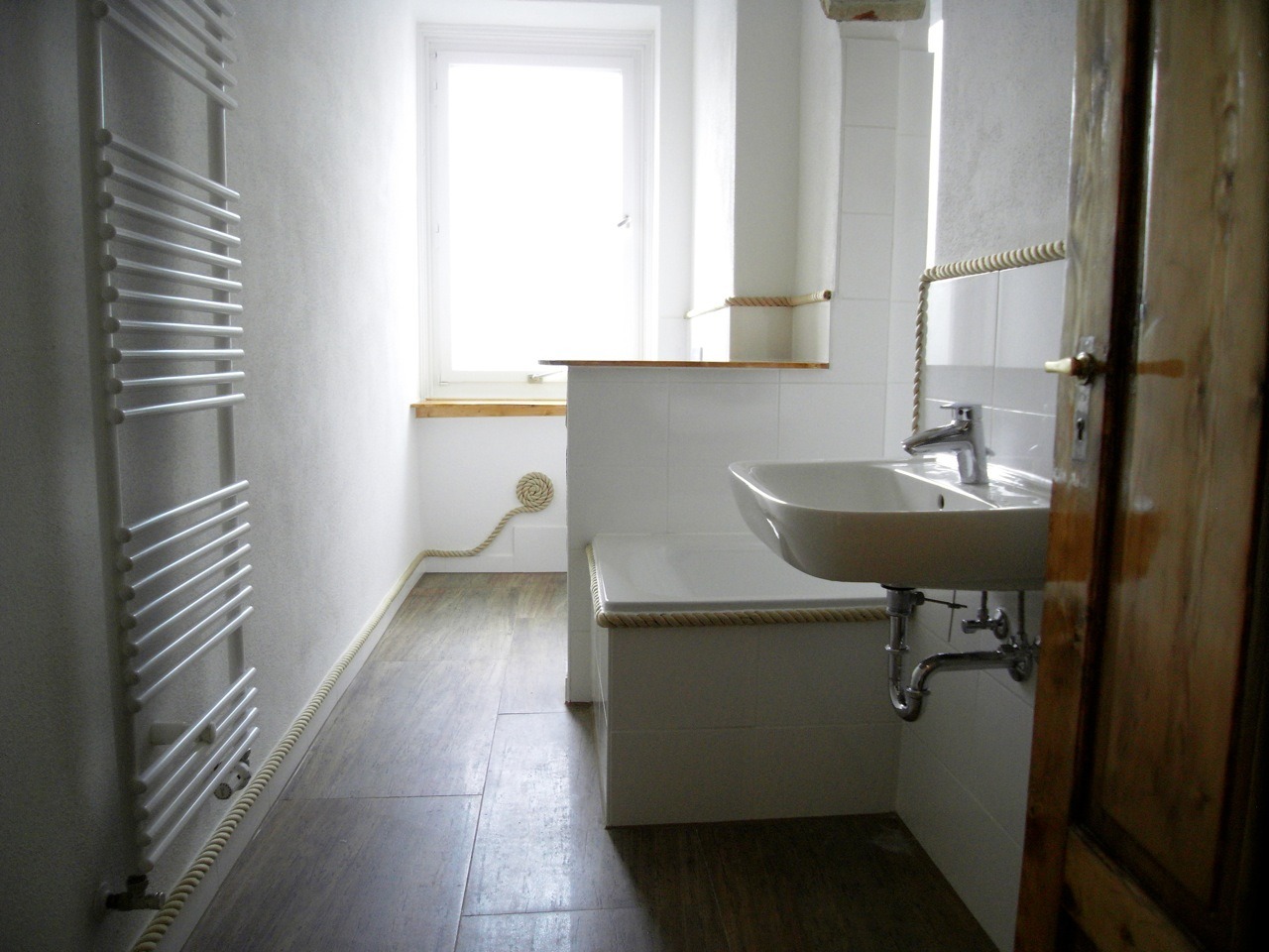 Jasna łazienka po remoncie z grzejnikiem drabinkowym, umywalką wiszącą i dekoracyjnym sznurem biegnącym wzdłuż ścian i wanny.