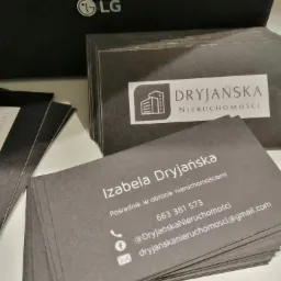 Stos wizytówek firmy Dryjańska Nieruchomości, z logo przedstawiającym budynek i danymi kontaktowymi pośrednika Izabeli Dryjańskiej, na tle czarnego pudełka z logo LG.